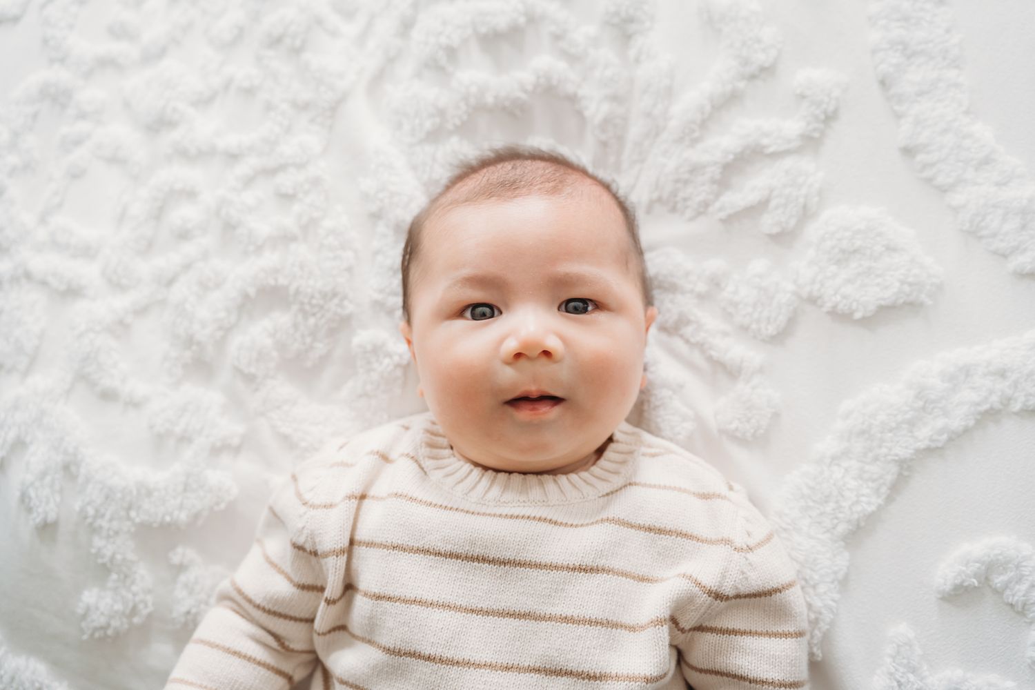 Baby wearing cream striped sweater lies on white textured background with sweet expression.