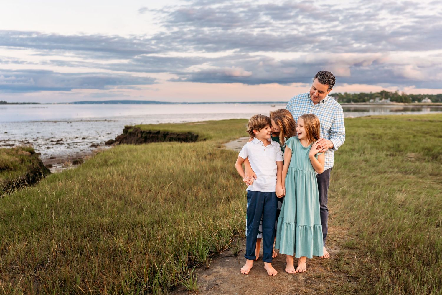 Scenic coastal landscape with tall grasses and cloudy skies along a beach at sunset.