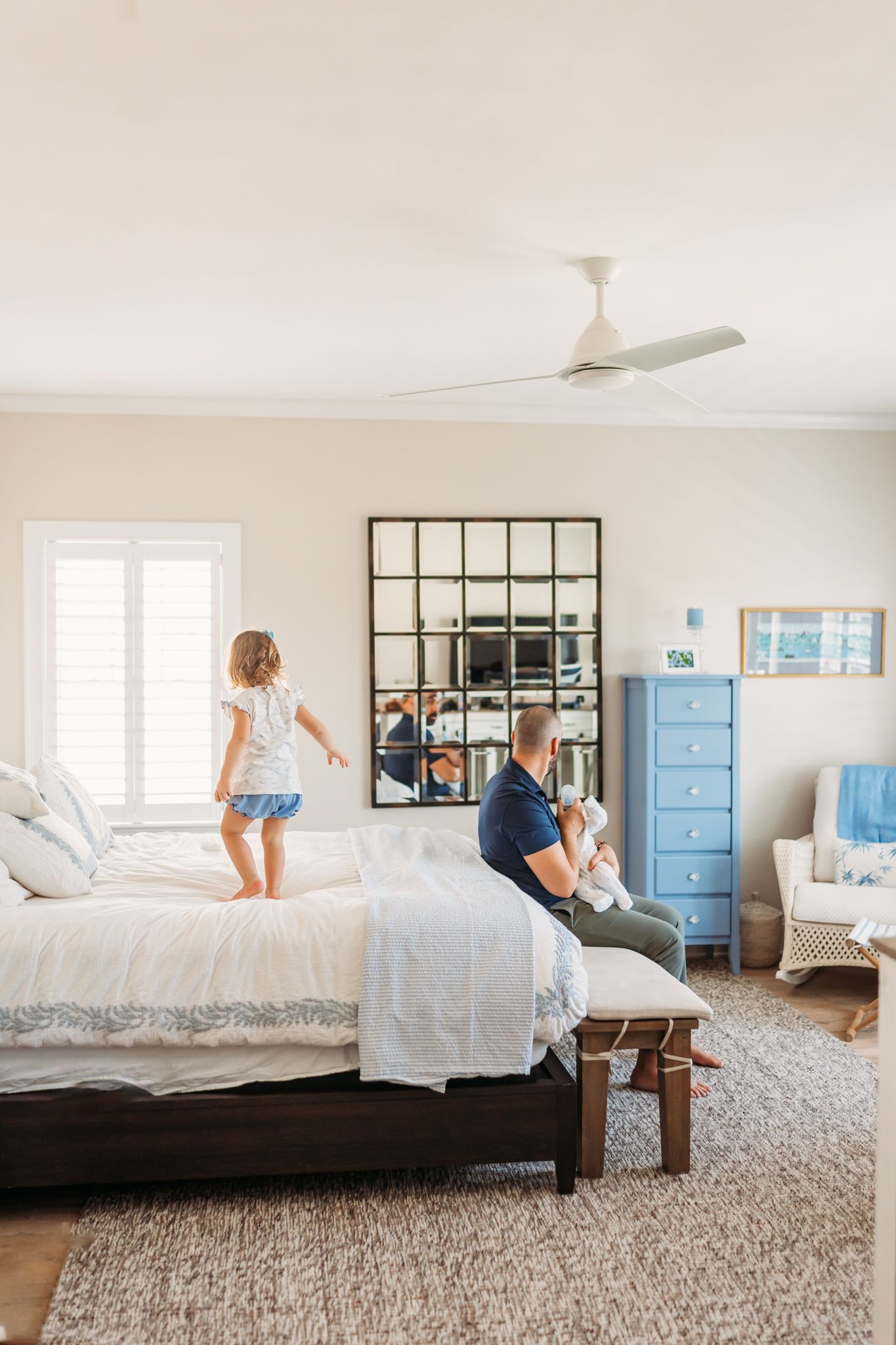 Light and airy bedroom with white bedding, blue dresser, and decorative mirror features playful activity on a comfortable bed.