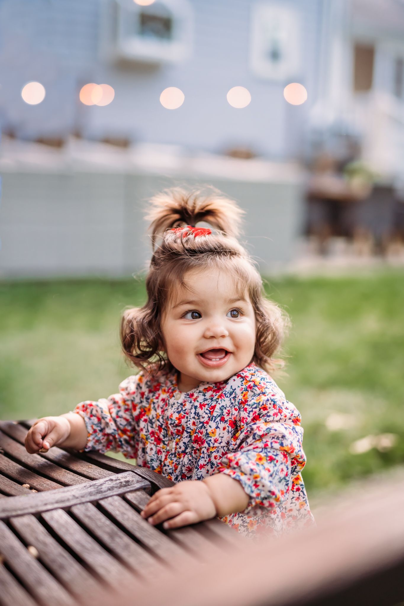 A toddler in a floral dress plays and smiles while leaning against a wooden bench outdoors.