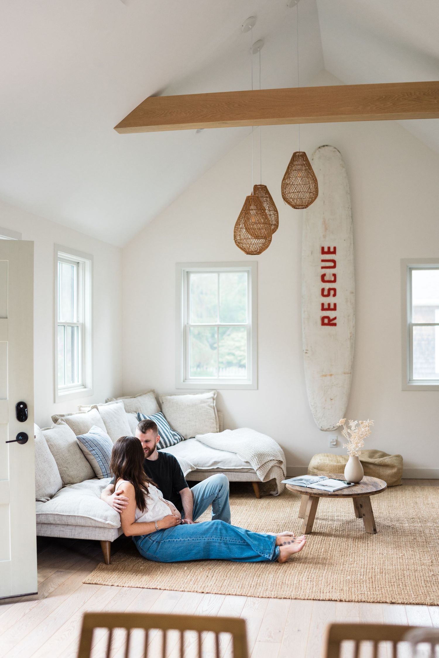 Cozy coastal living room with rattan pendant lights, surfboard decor, and natural jute rug on white wood floors.