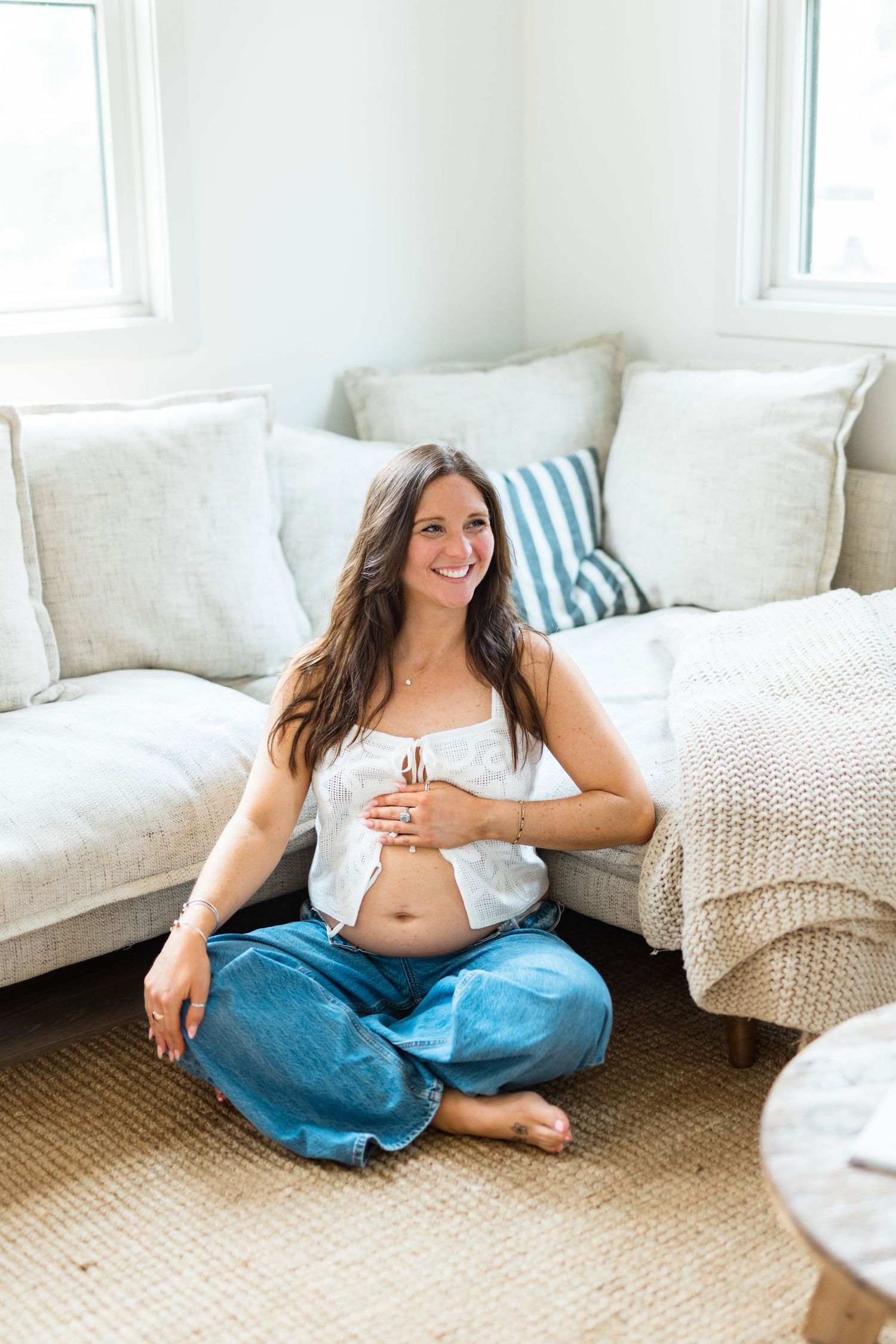 A woman in blue jeans and white top relaxes casually on a beige carpet beside a white sofa in a bright room.