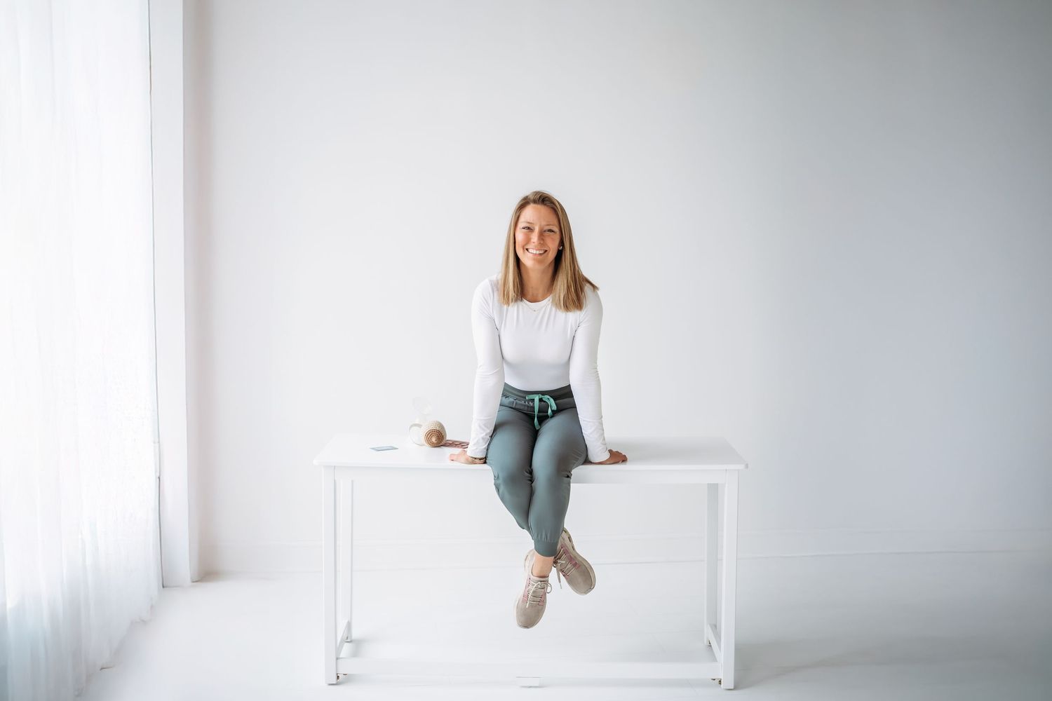 Woman in casual attire sits and poses on white furniture against a light background in a minimalist setting.