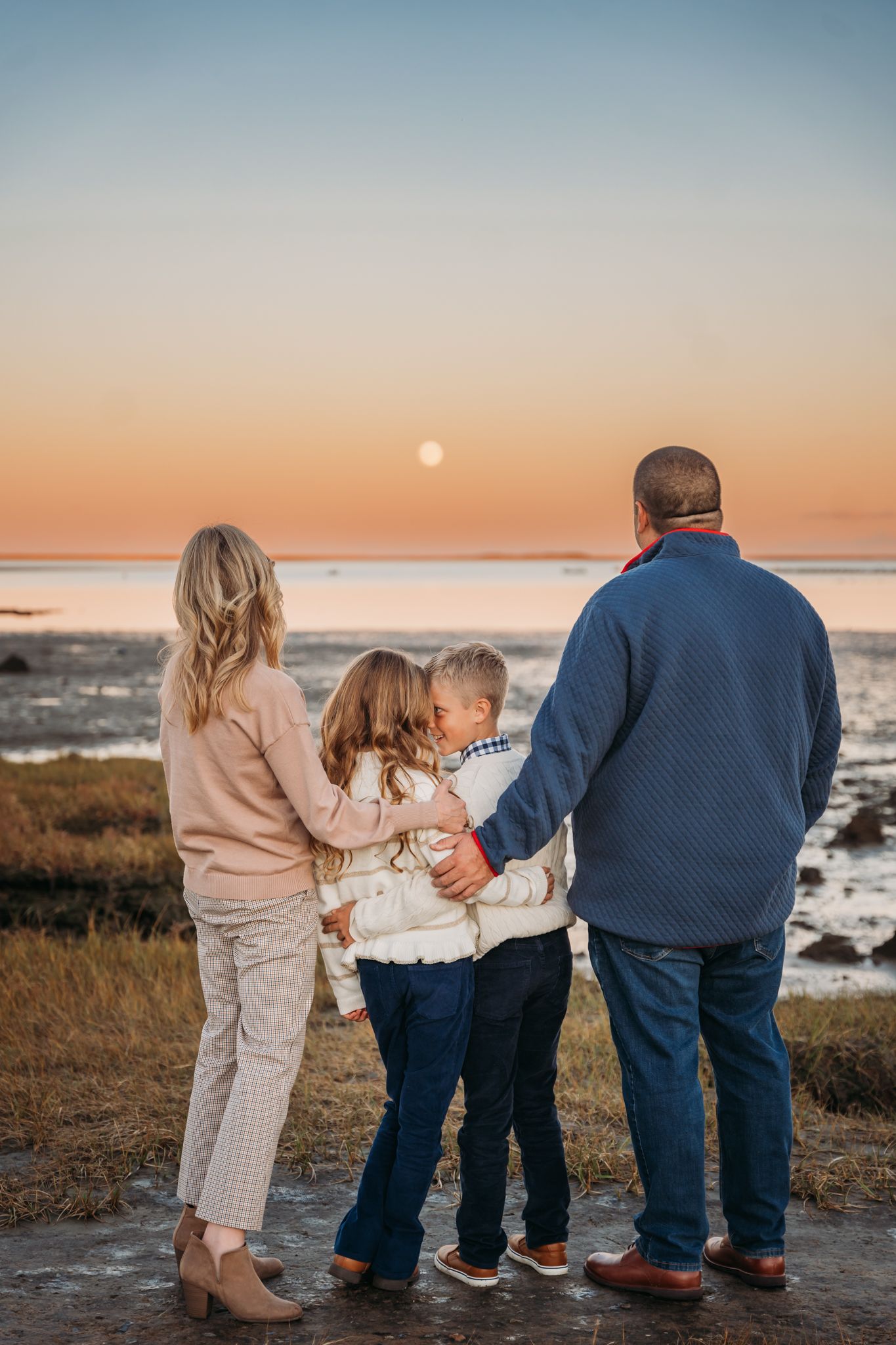A family stands together watching the sunset over the ocean while the full moon rises in the pastel sky.
