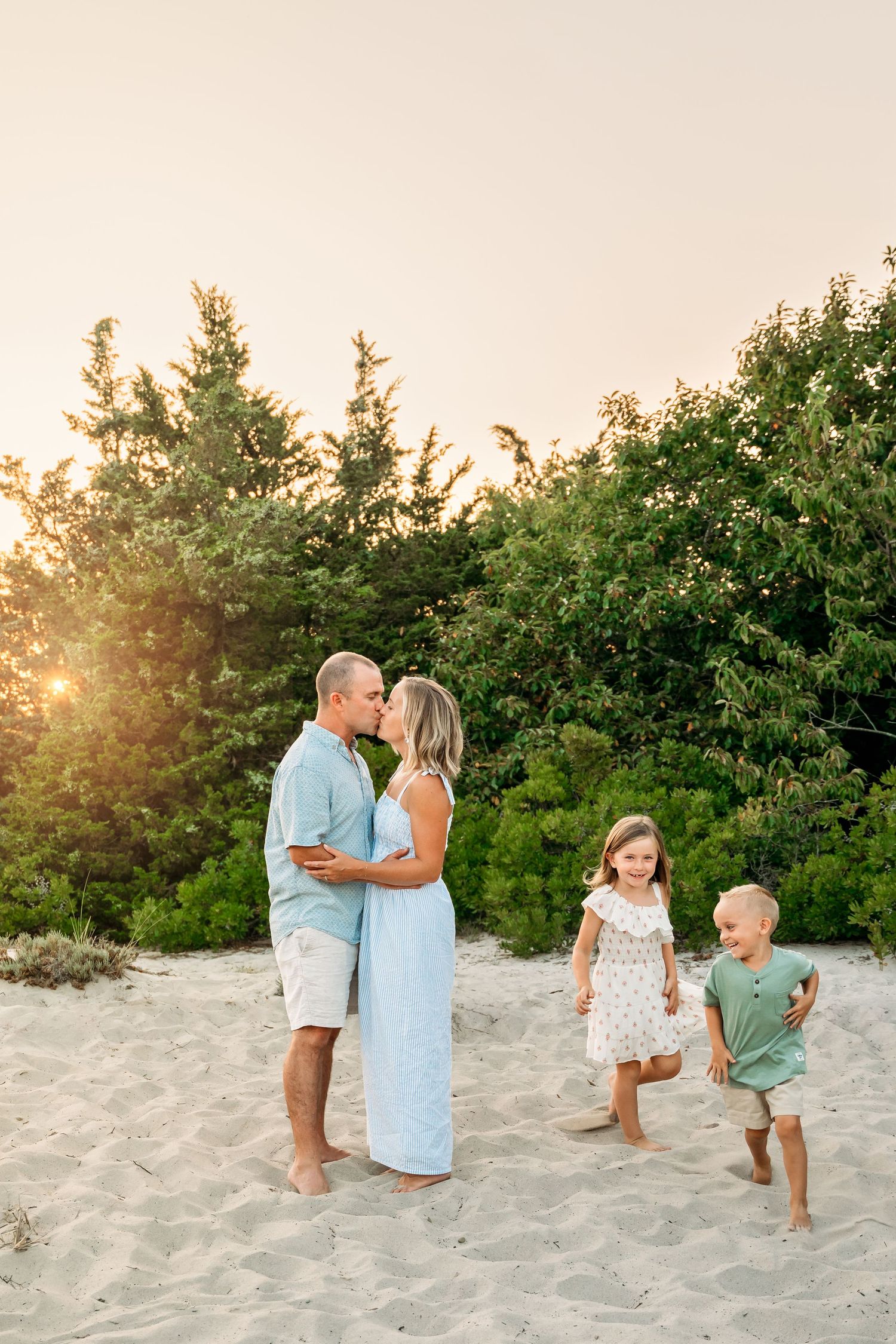 A loving family enjoys a sunset photoshoot together on a sandy beach with green foliage in the background.