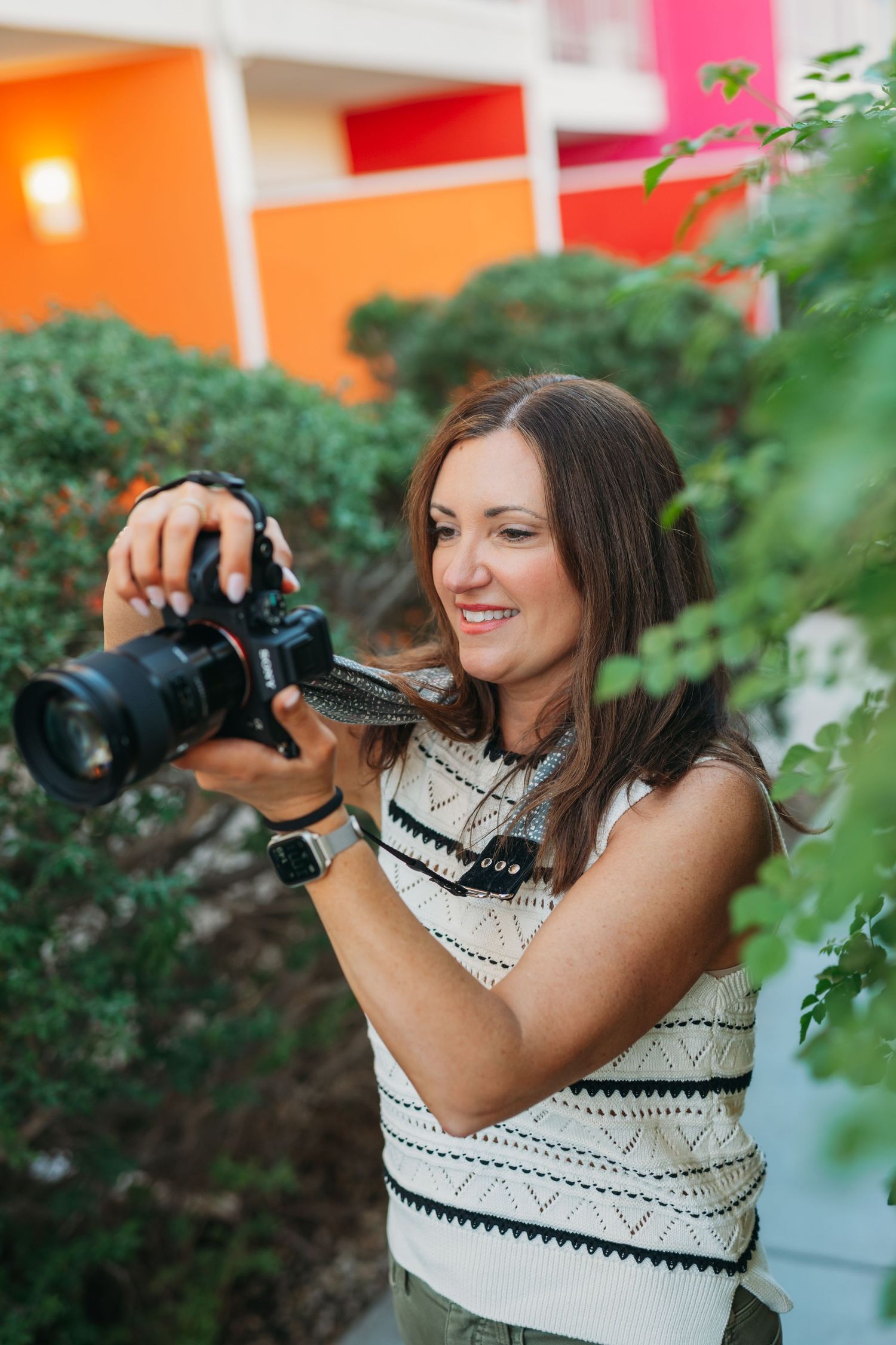 A photographer in a white crochet top captures photos through green foliage in a vibrant orange and red setting.