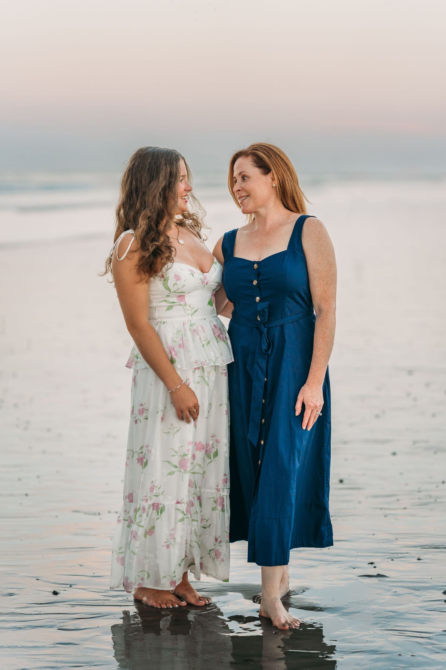 Two women in summer dresses share a moment together on a beach at sunset, standing in shallow water.