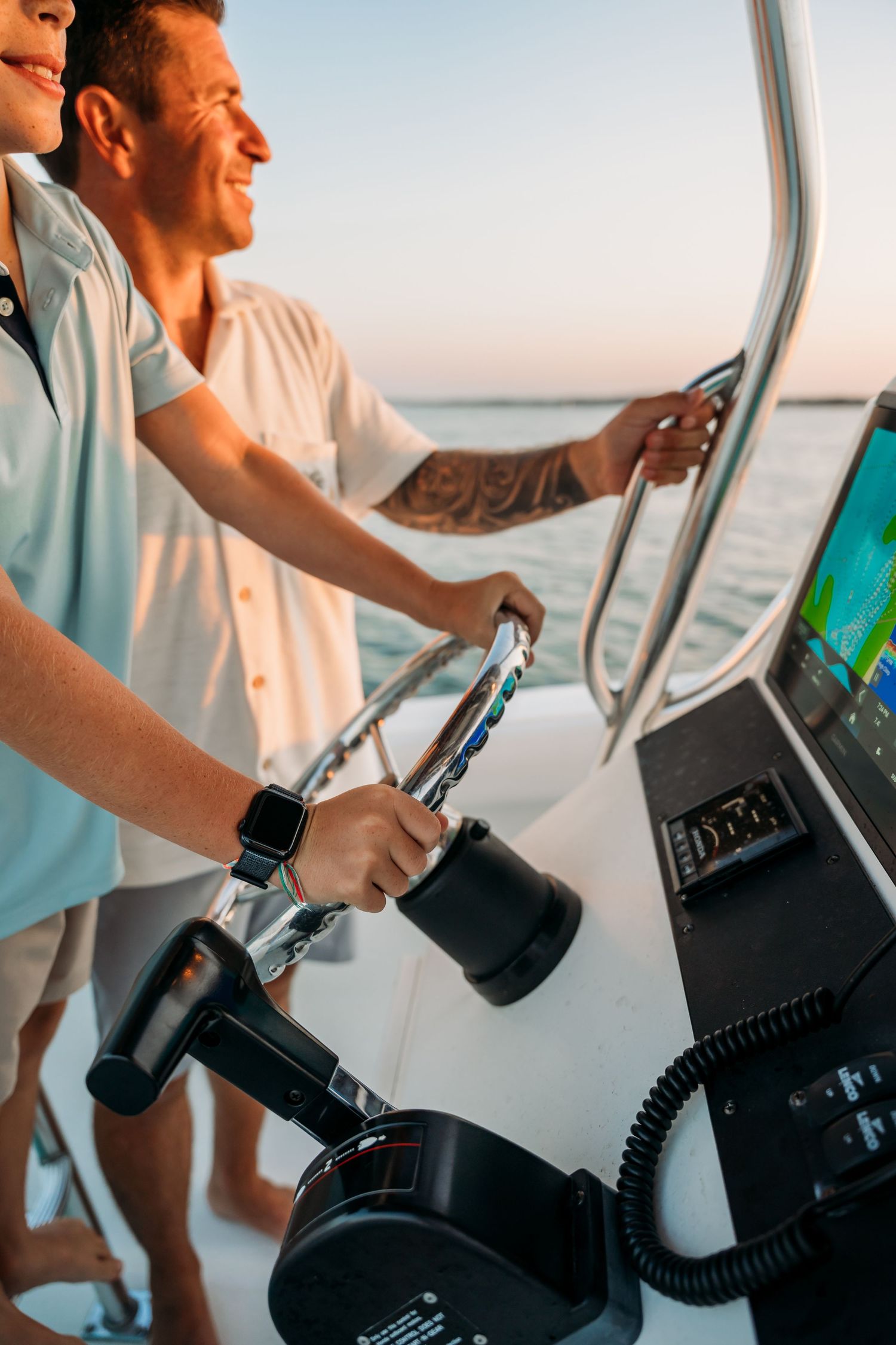 A person's hand grips a steering wheel on a boat during a scenic sunset cruise.