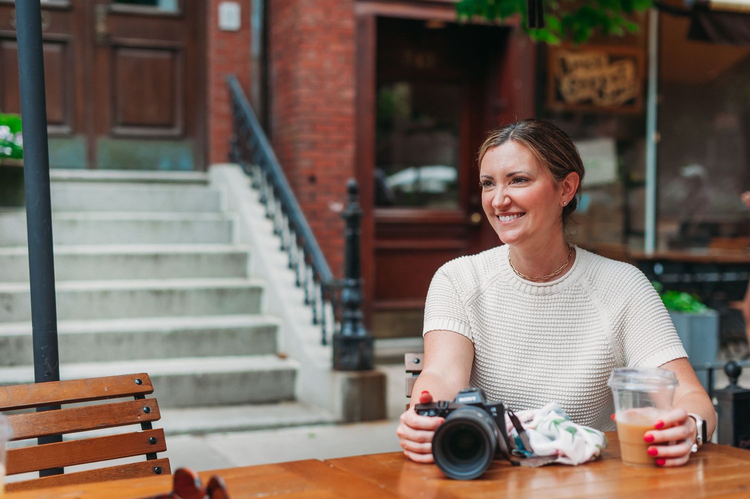 Two people sit at an outdoor cafe table with drinks and a camera, enjoying a sunny afternoon conversation.