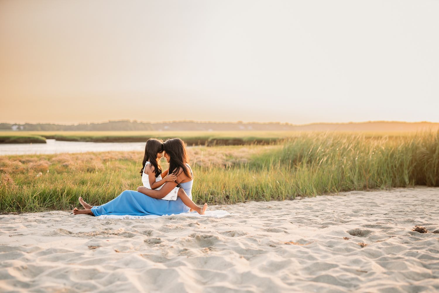 Two women sit closely together on a sandy beach at golden hour, surrounded by tall green marsh grass.