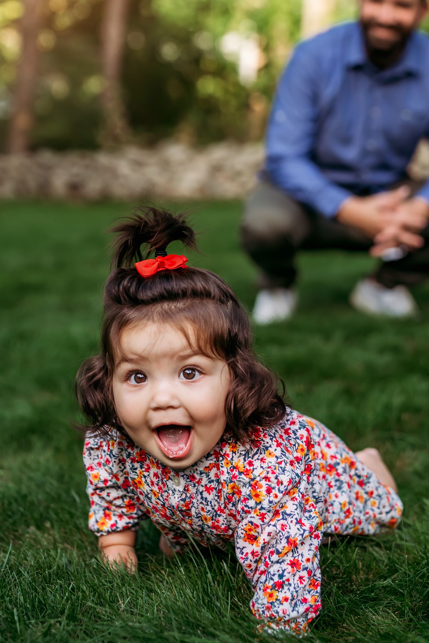 A small child in a floral print dress crawls and plays on green grass during a sunny outdoor photo session.