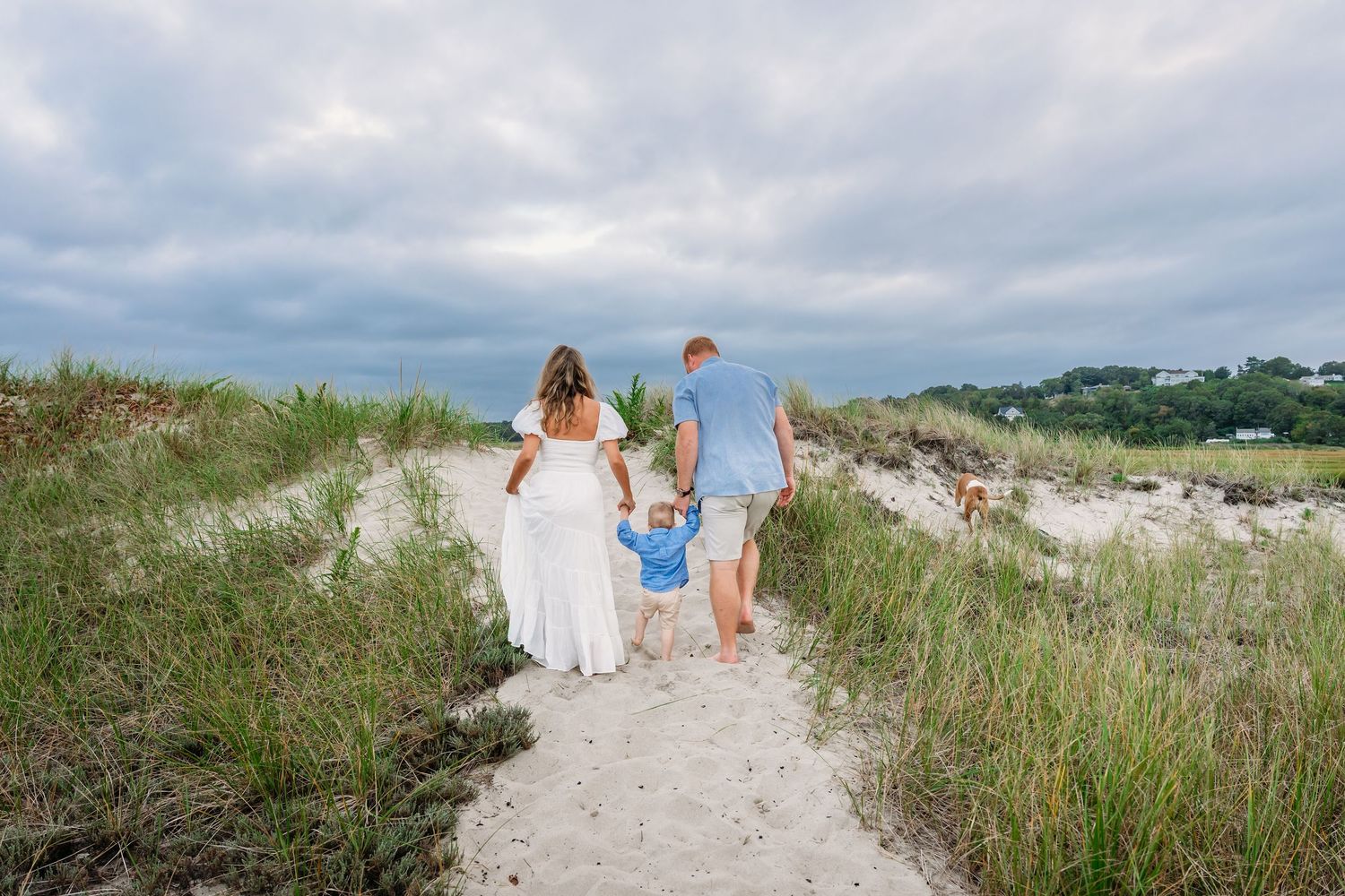 Family of three walking hand-in-hand on a sandy beach path surrounded by dunes and green grass under cloudy skies.