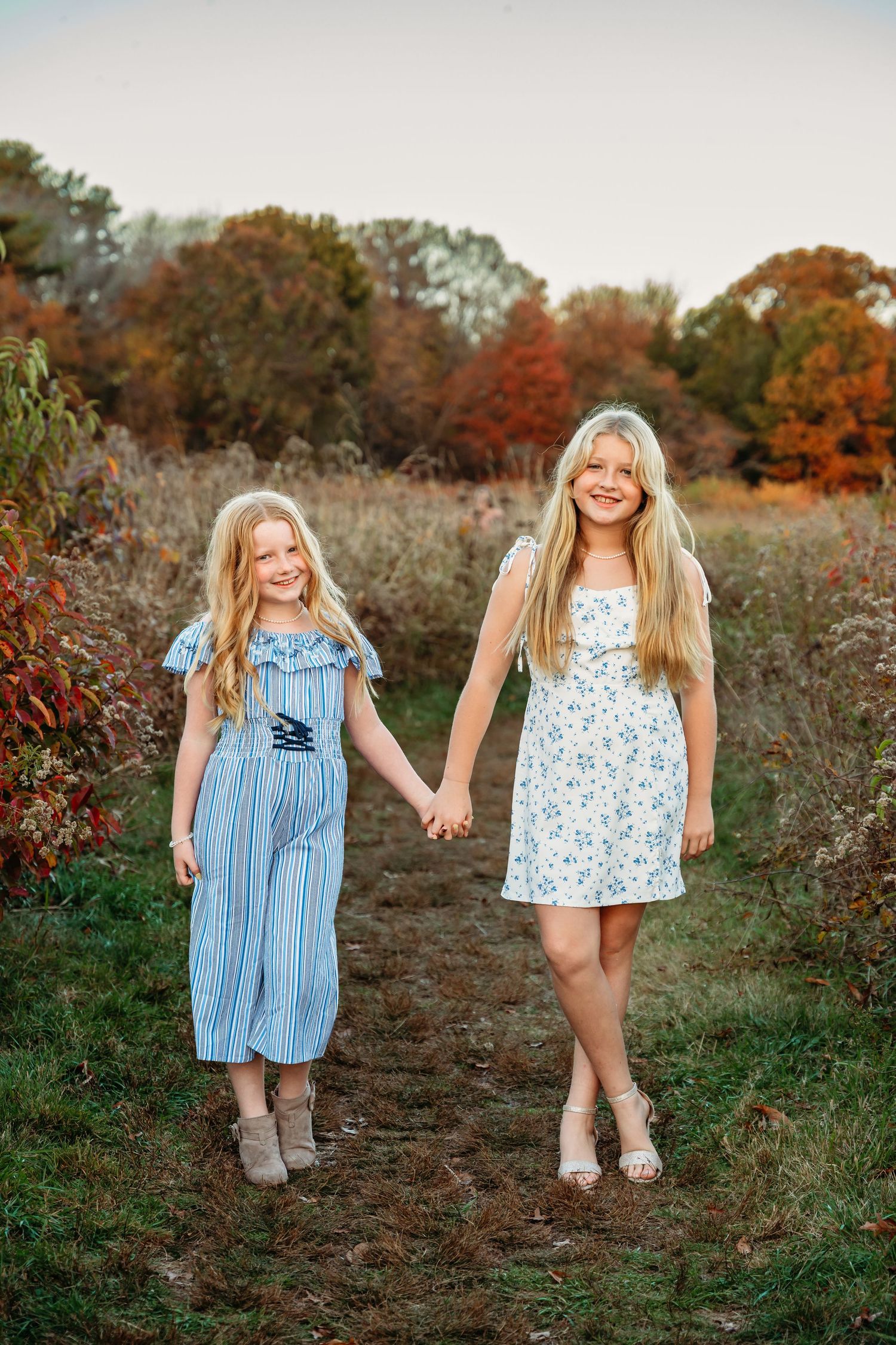 Two children in blue and floral dresses play together in a field with autumn foliage in the background.