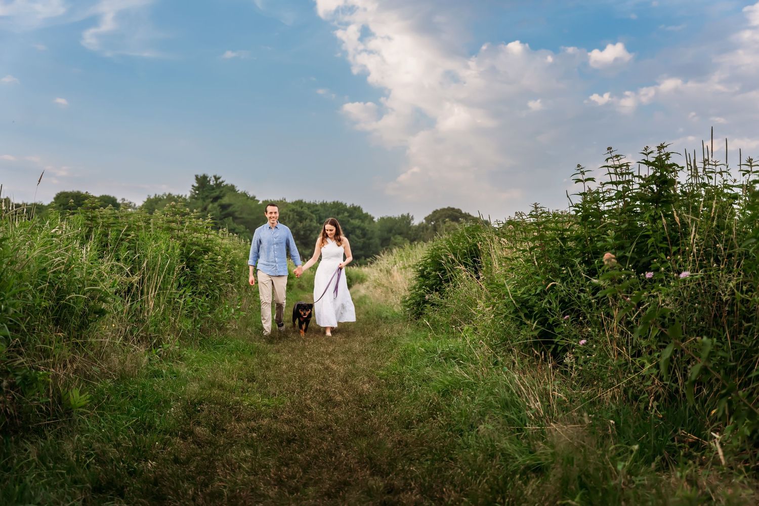 A couple walks through a lush green meadow on a sunny summer day with puffy white clouds in the sky.