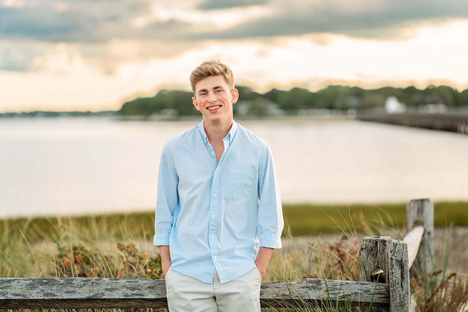 Young man in light blue shirt and khaki pants smiling outdoors near a waterfront with a wooden fence.