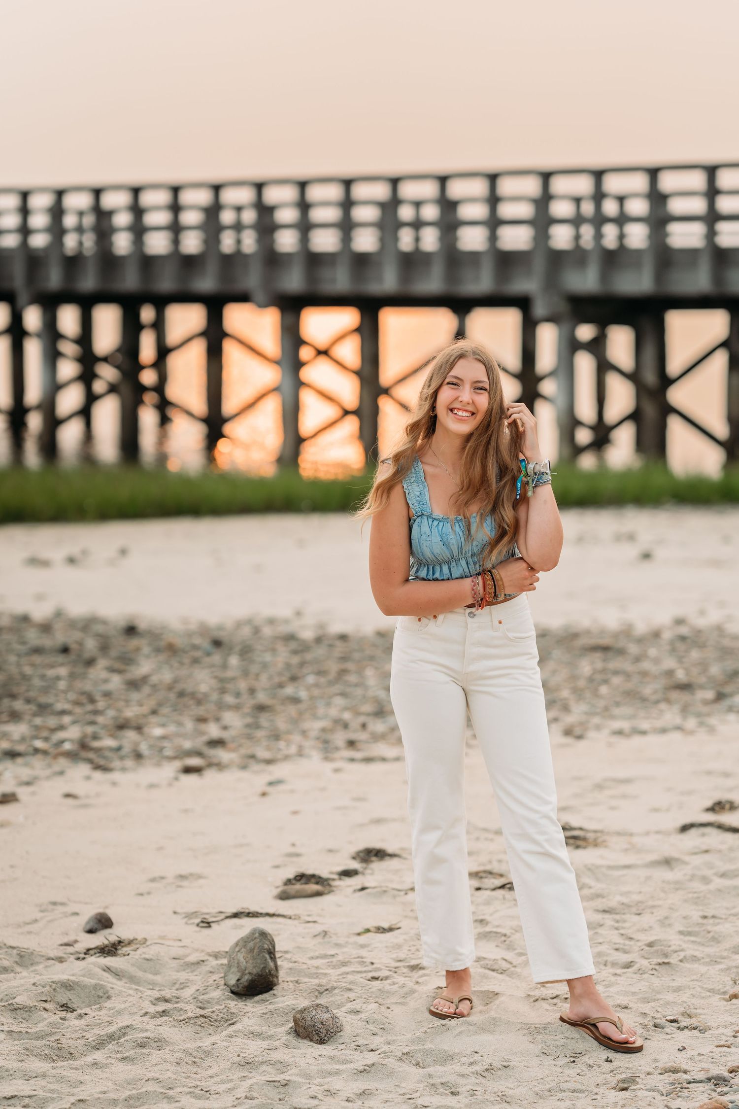 Young woman in white jeans and blue top smiling on sandy beach at sunset with wooden pier in background.