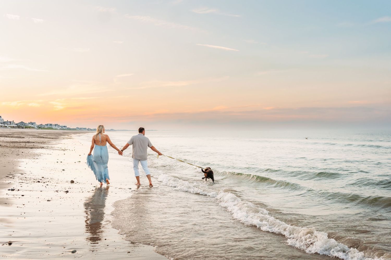 A couple walking their dog along a serene beach shoreline during a beautiful golden sunset.