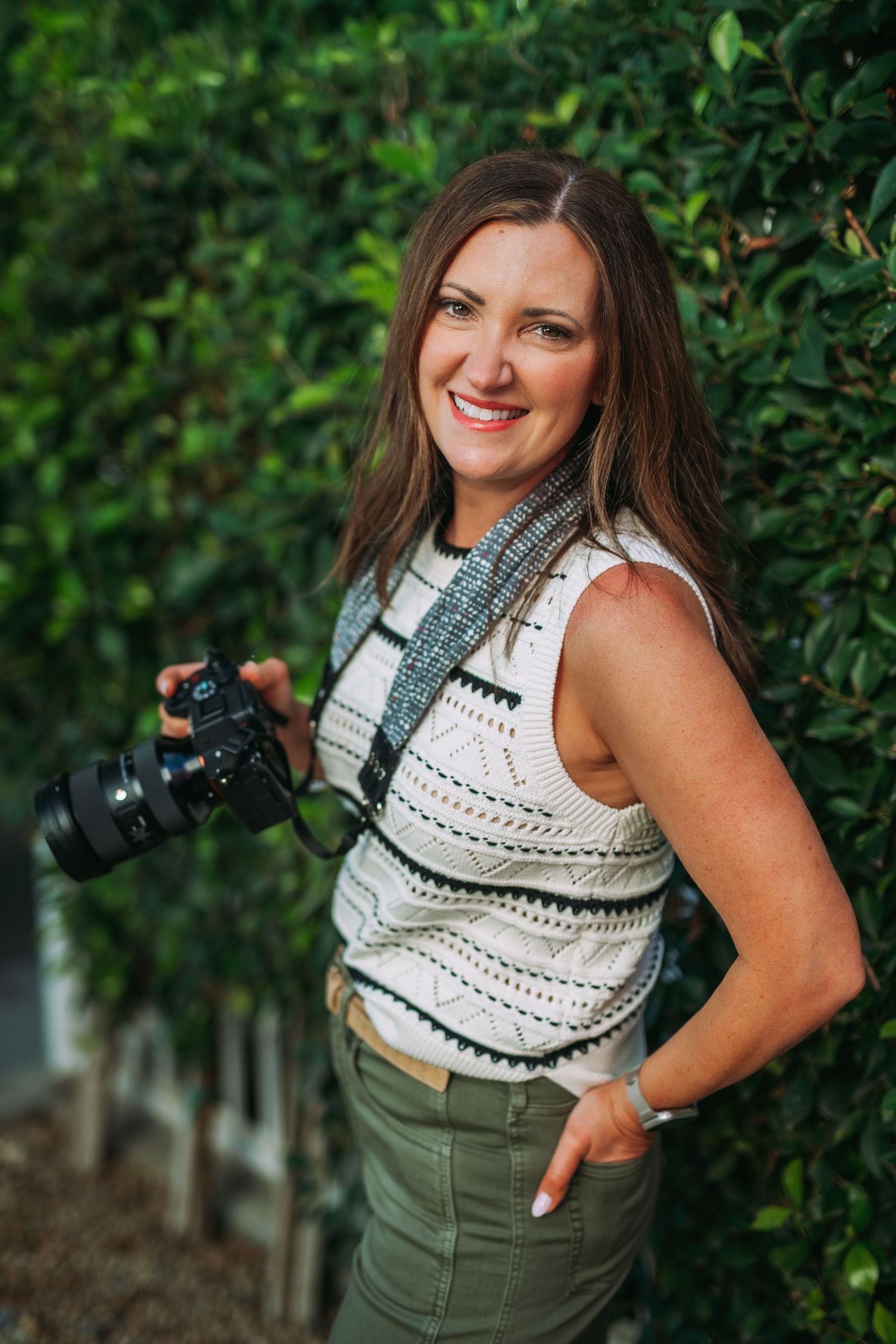 A photographer in a white sleeveless top and olive pants poses with a camera against a leafy green backdrop.