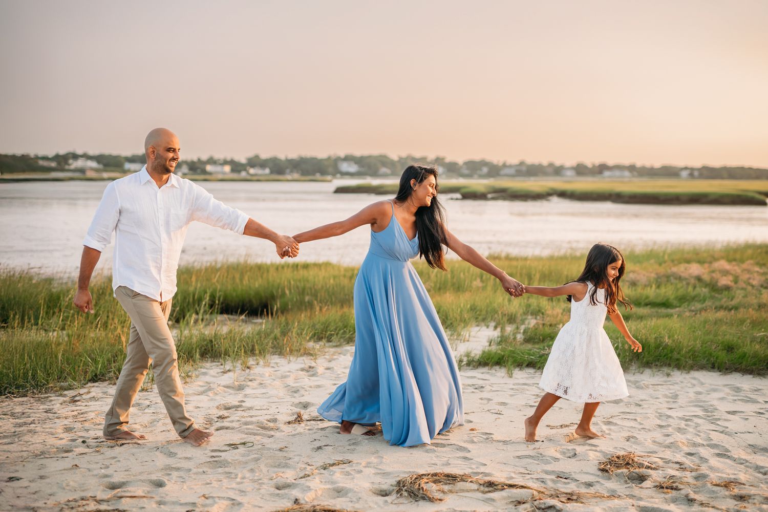 Family of three holding hands walking on a sandy beach at sunset, wearing white and blue outfits.