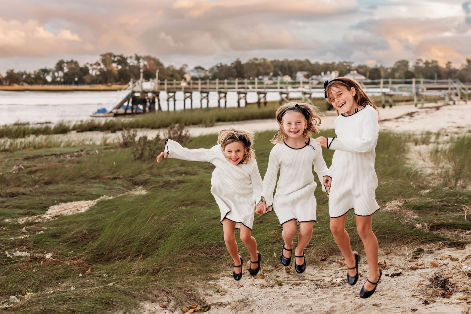 Three children in white outfits jump and play together on a grassy coastal area during sunset.