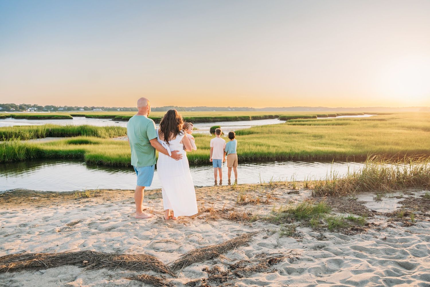 A family walks along a scenic coastal beach path during golden hour sunset with water and marsh views.