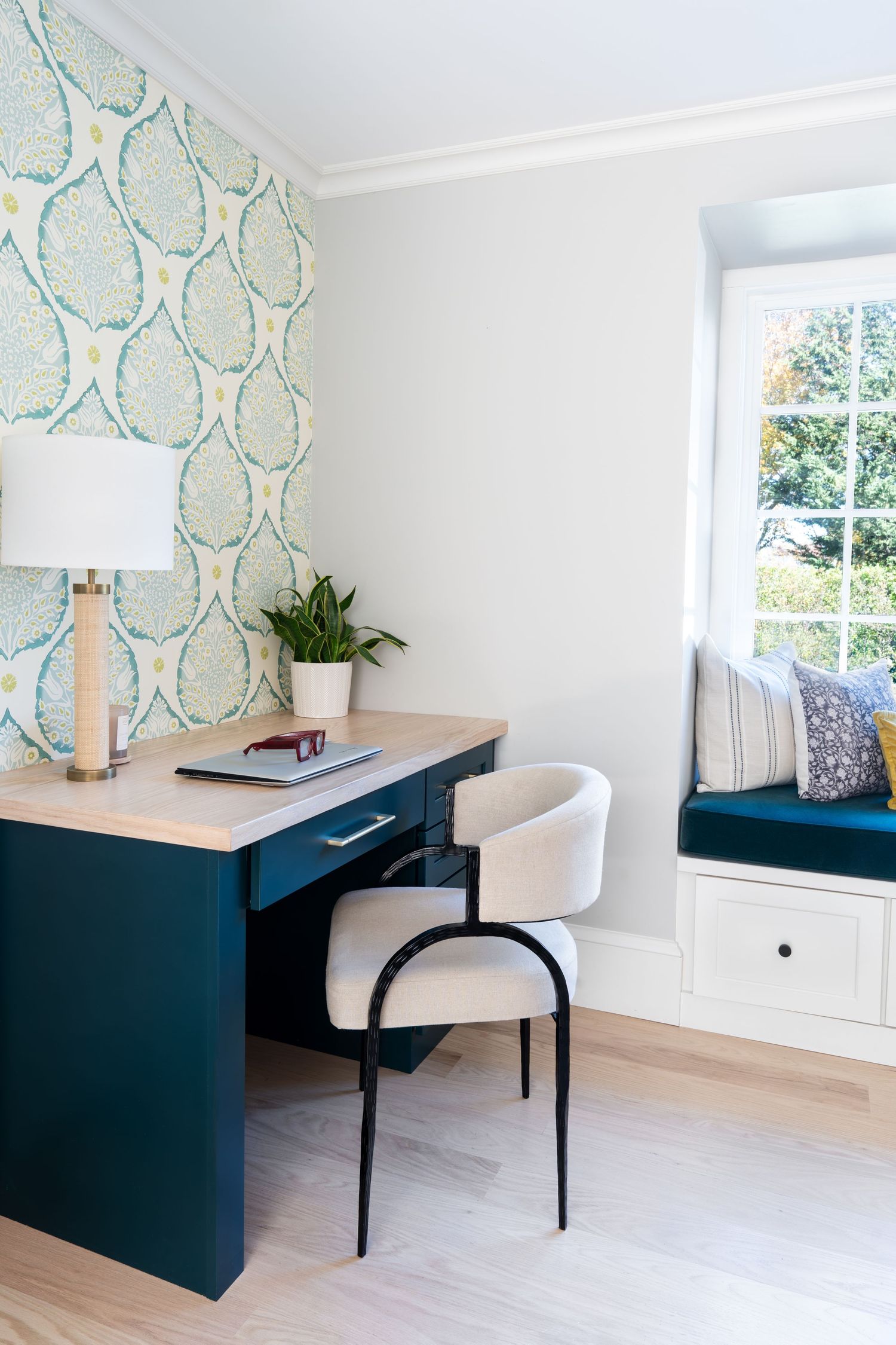 Modern home office features navy blue built-in desk, white upholstered chair, and blue botanical wallpaper in bright space.
