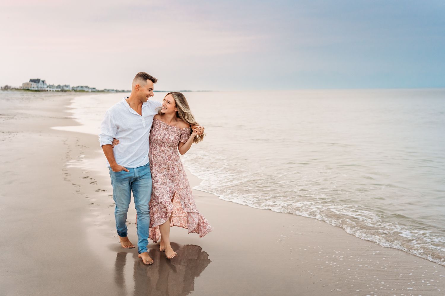 A couple in casual attire playfully walks along a sunny beach with waves rolling in behind them.