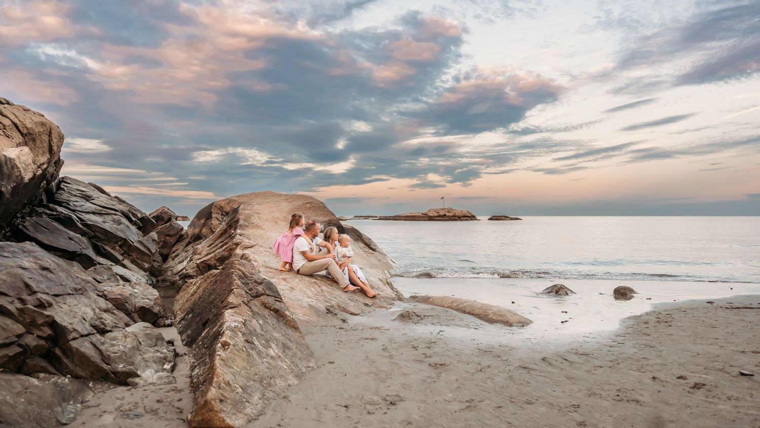 Family sitting on coastal rocks during dramatic sunset with pink and blue clouds at Minot Beach.