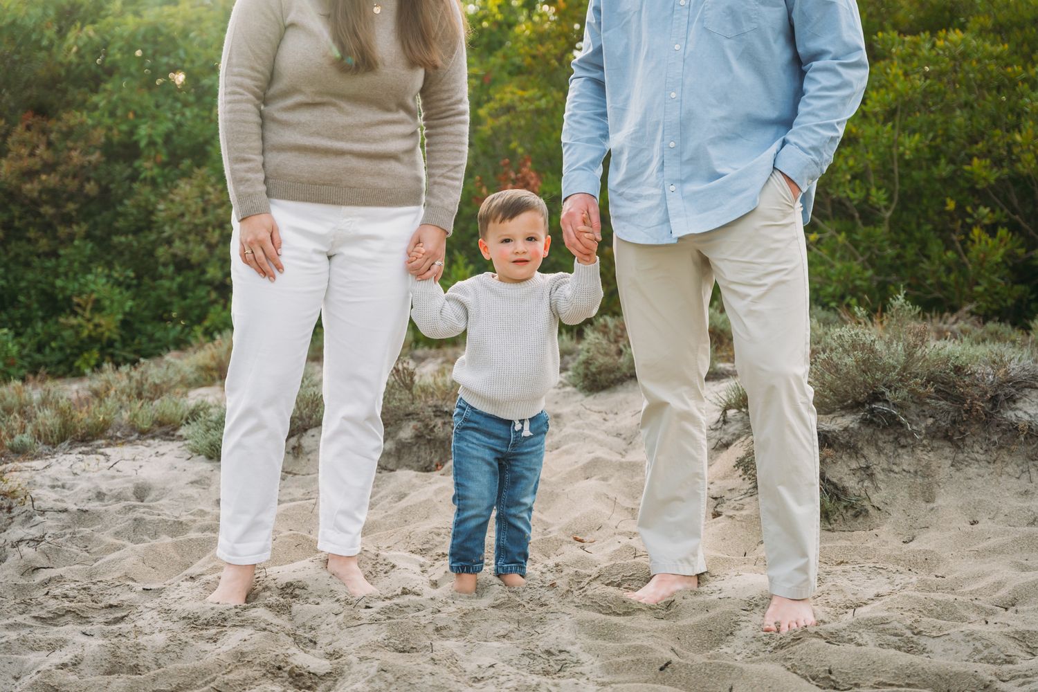 Family of three standing barefoot on sandy beach, parents holding toddler's hands outdoors.