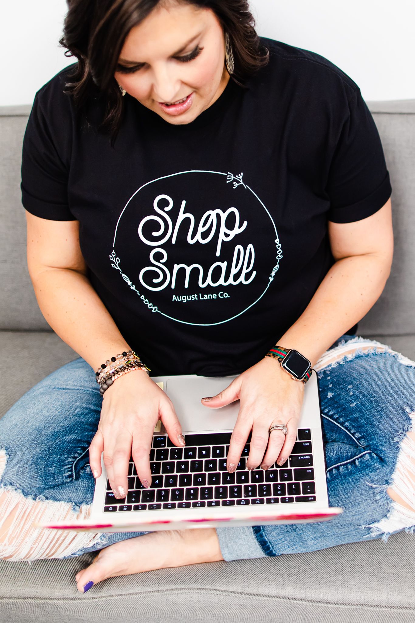 Person wearing black Shop t-shirt and jeans sits cross-legged while working on laptop computer.