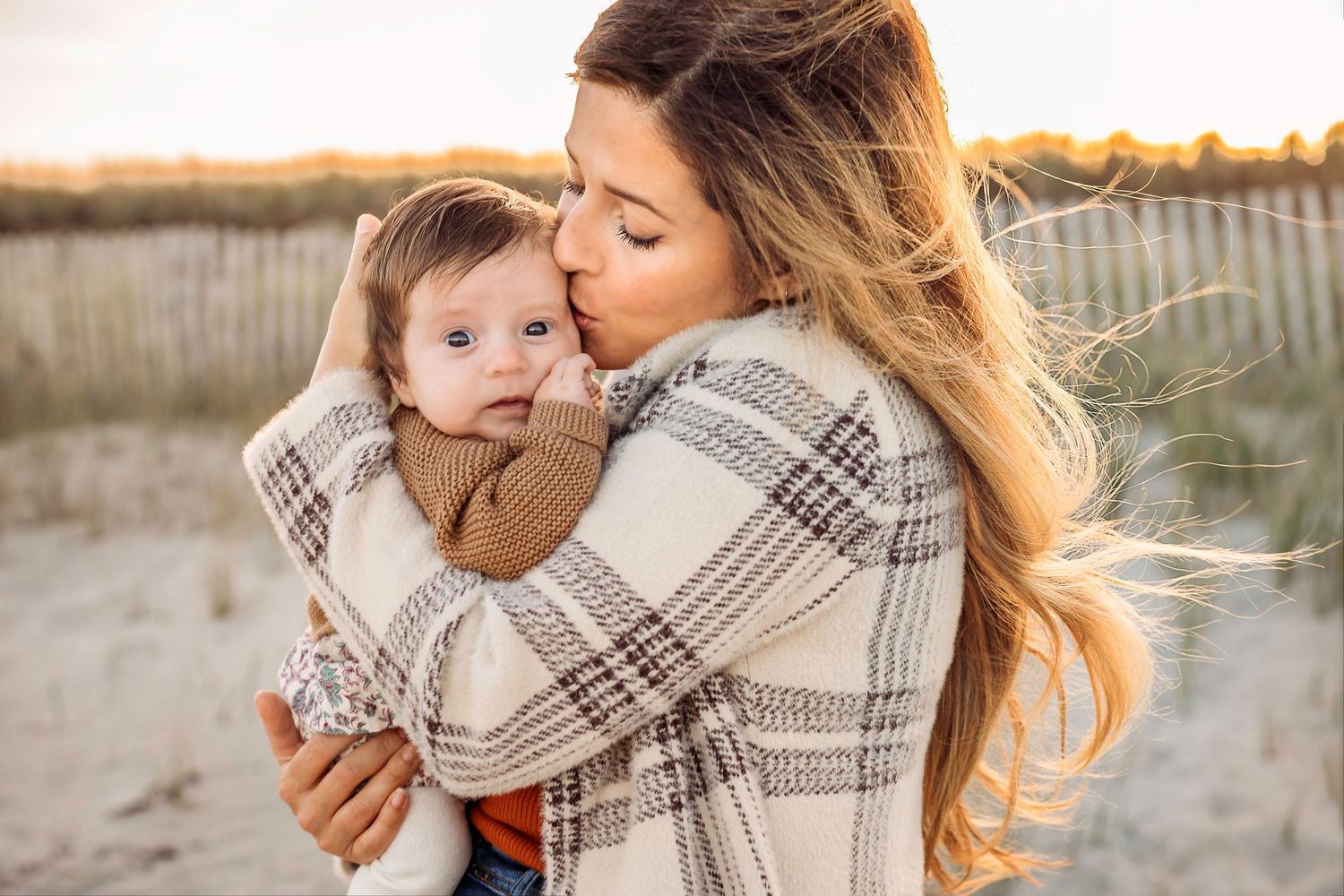 Mother wrapped in plaid blanket holding small child on Duxbury Beach at sunset.