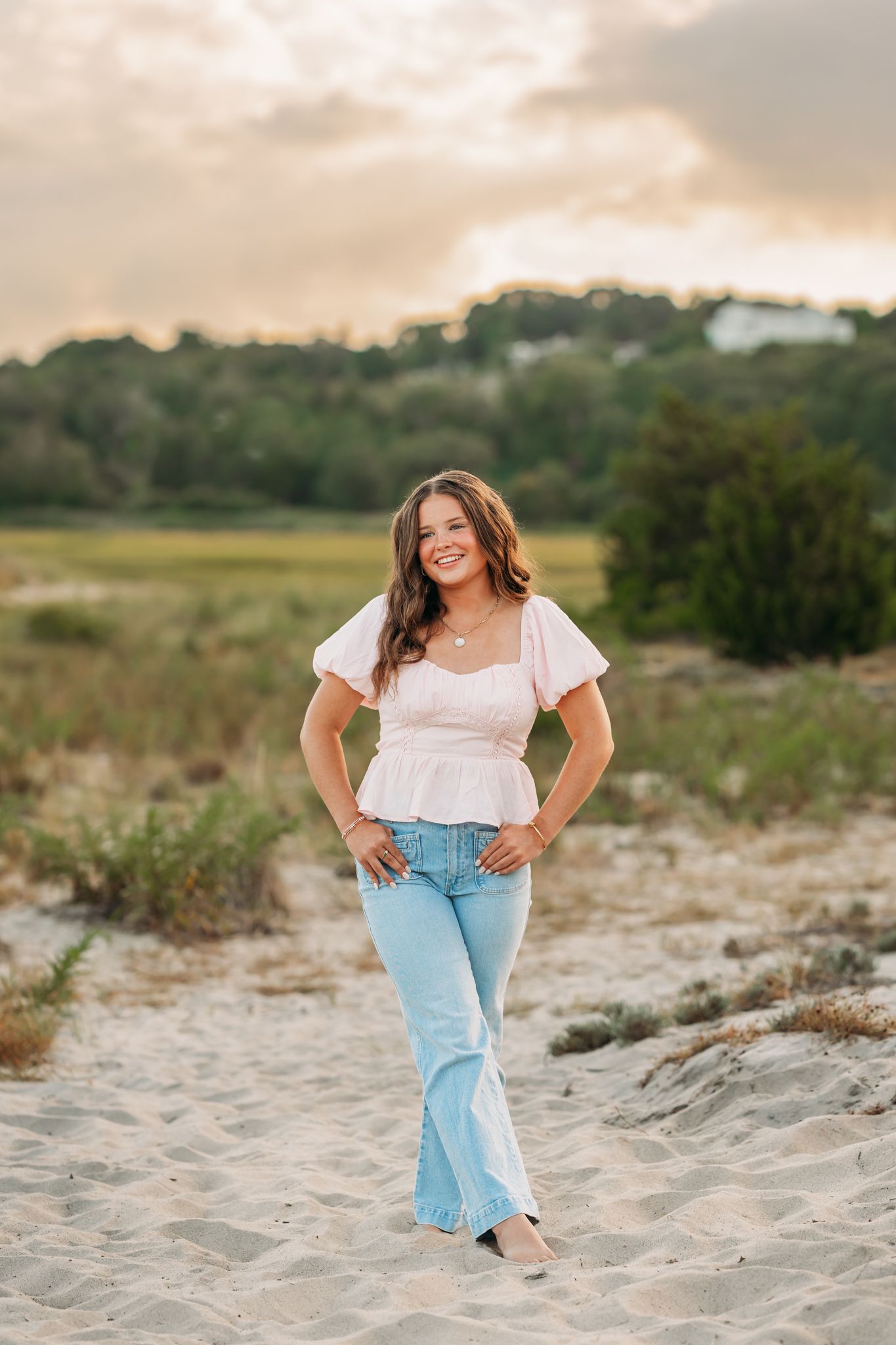Young woman in pink top and flare jeans posing on sandy beach path at golden hour sunset.