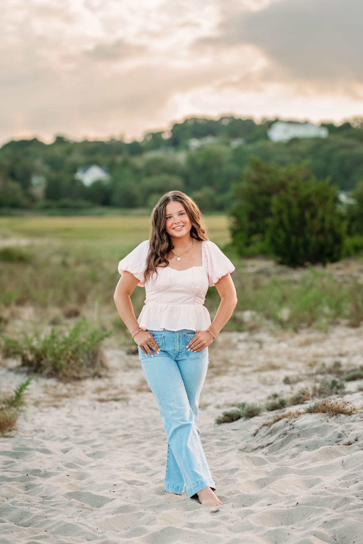 Young woman in pink top and flare jeans posing on sandy beach with green trees and cloudy sky backdrop.