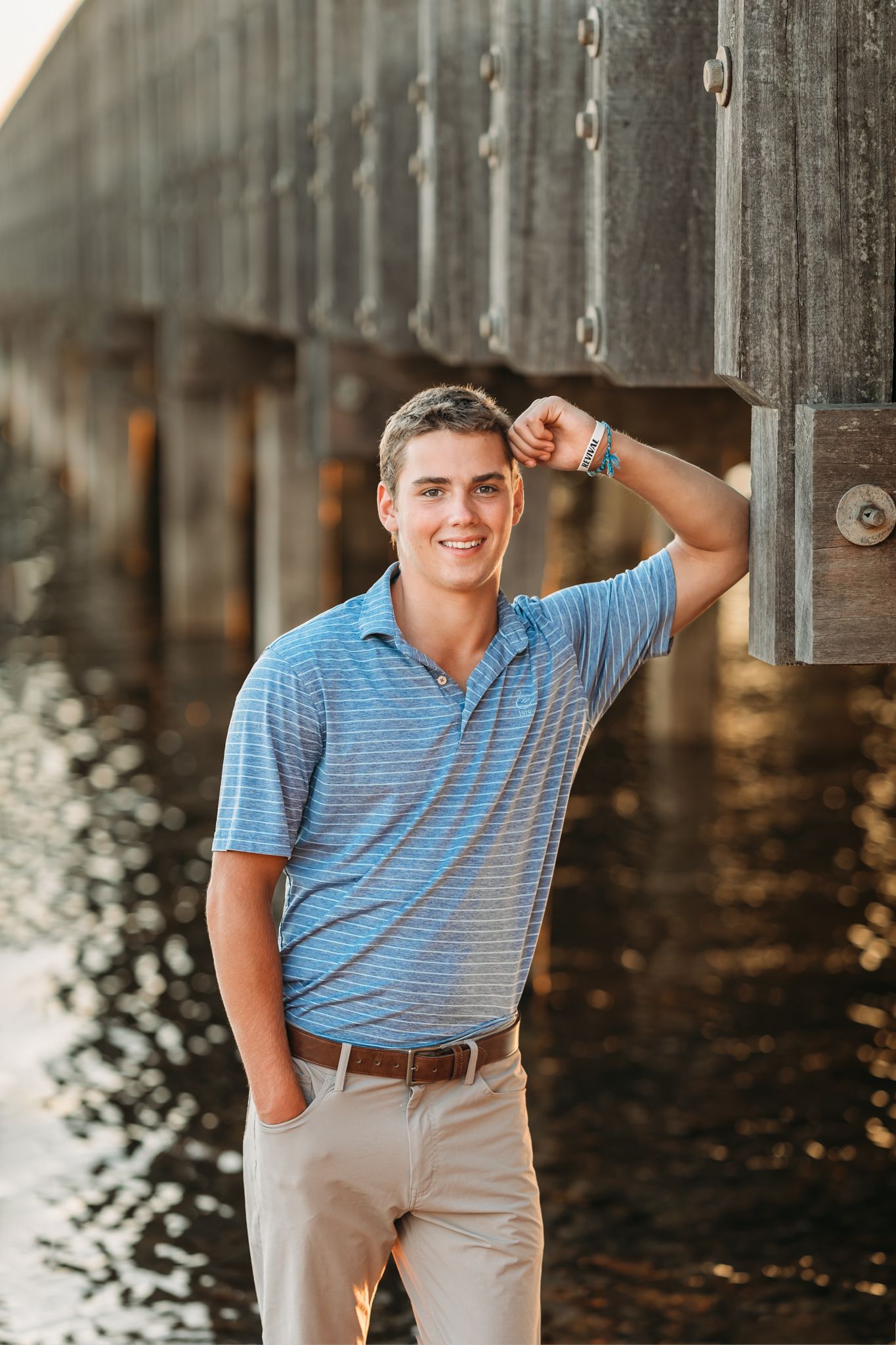 A smiling young man in a blue polo shirt poses confidently by a waterfront pier in golden hour light.