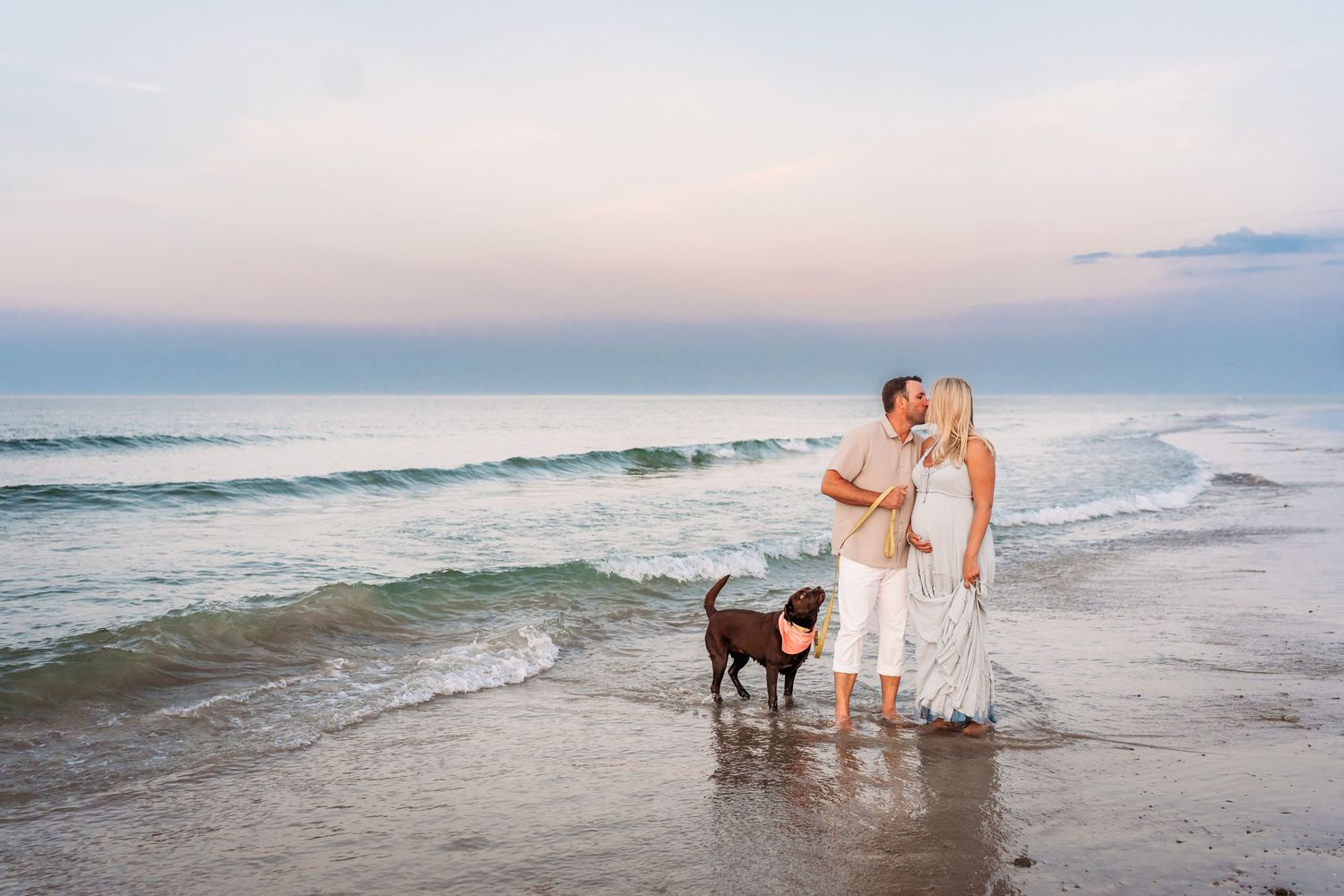 Couple expecting a child standing in ocean waves with dog during pastel sunset. Black lab dog is looking up at them.