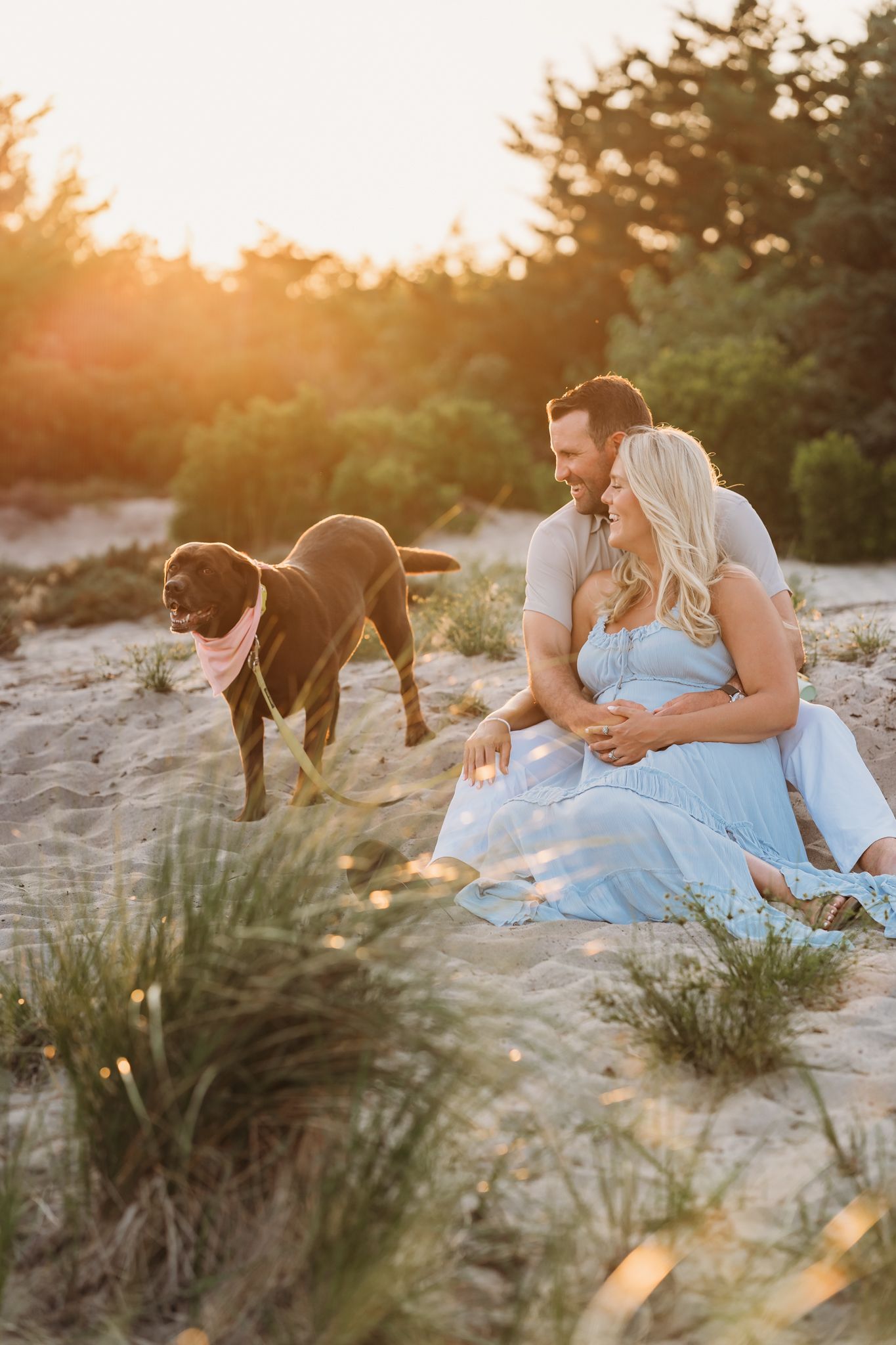 A couple sits together on sandy dunes with their brown dog during a golden sunset, surrounded by beach grass.