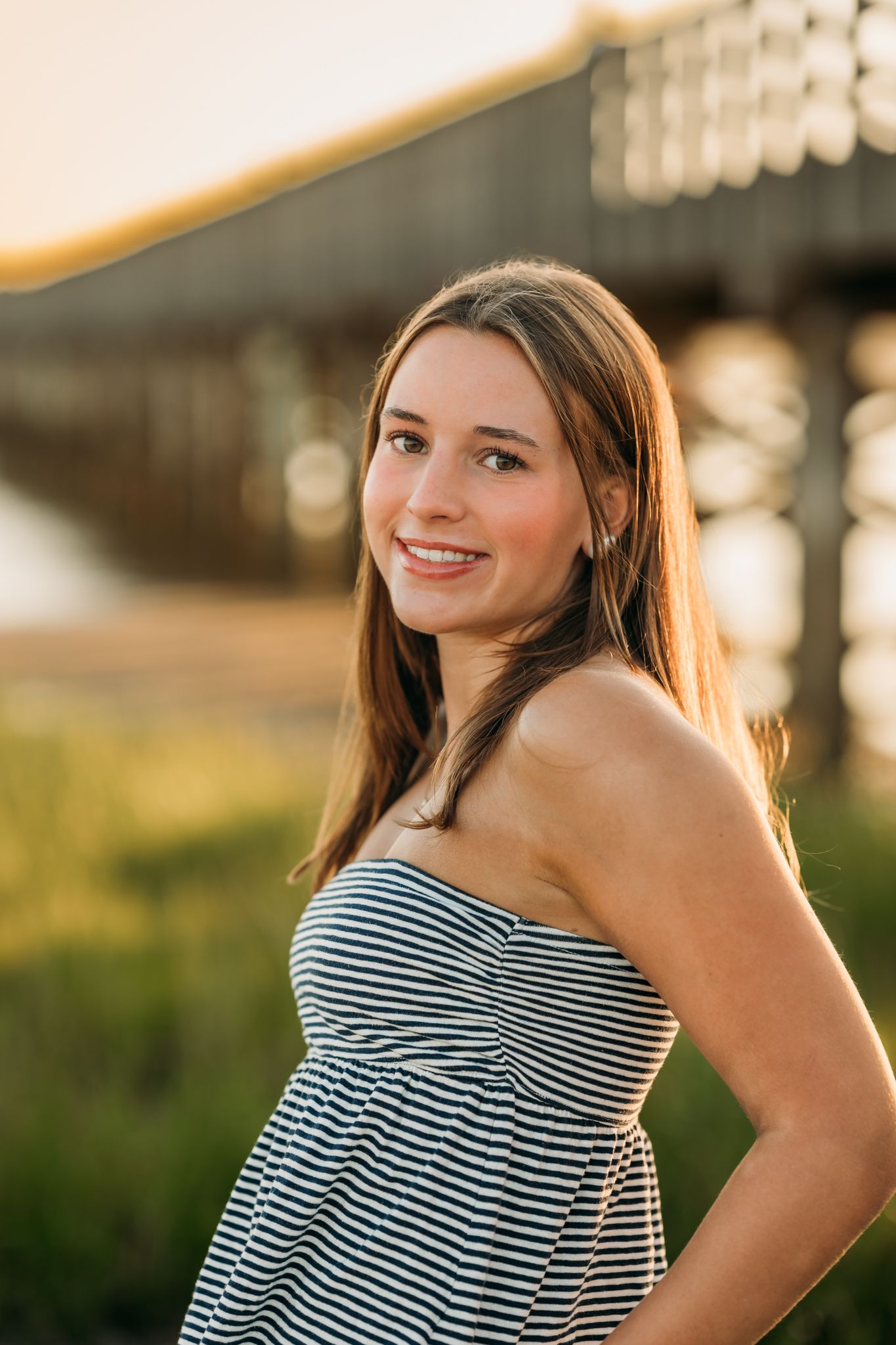Young woman in striped strapless dress smiling outdoors near a pier during golden hour sunset.