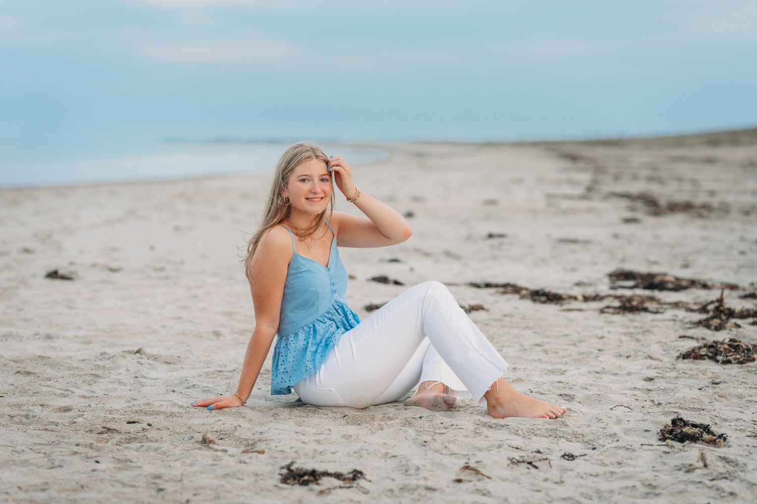 Young blonde woman in blue top and white jeans sitting barefoot on a sandy beach with calm ocean in background.