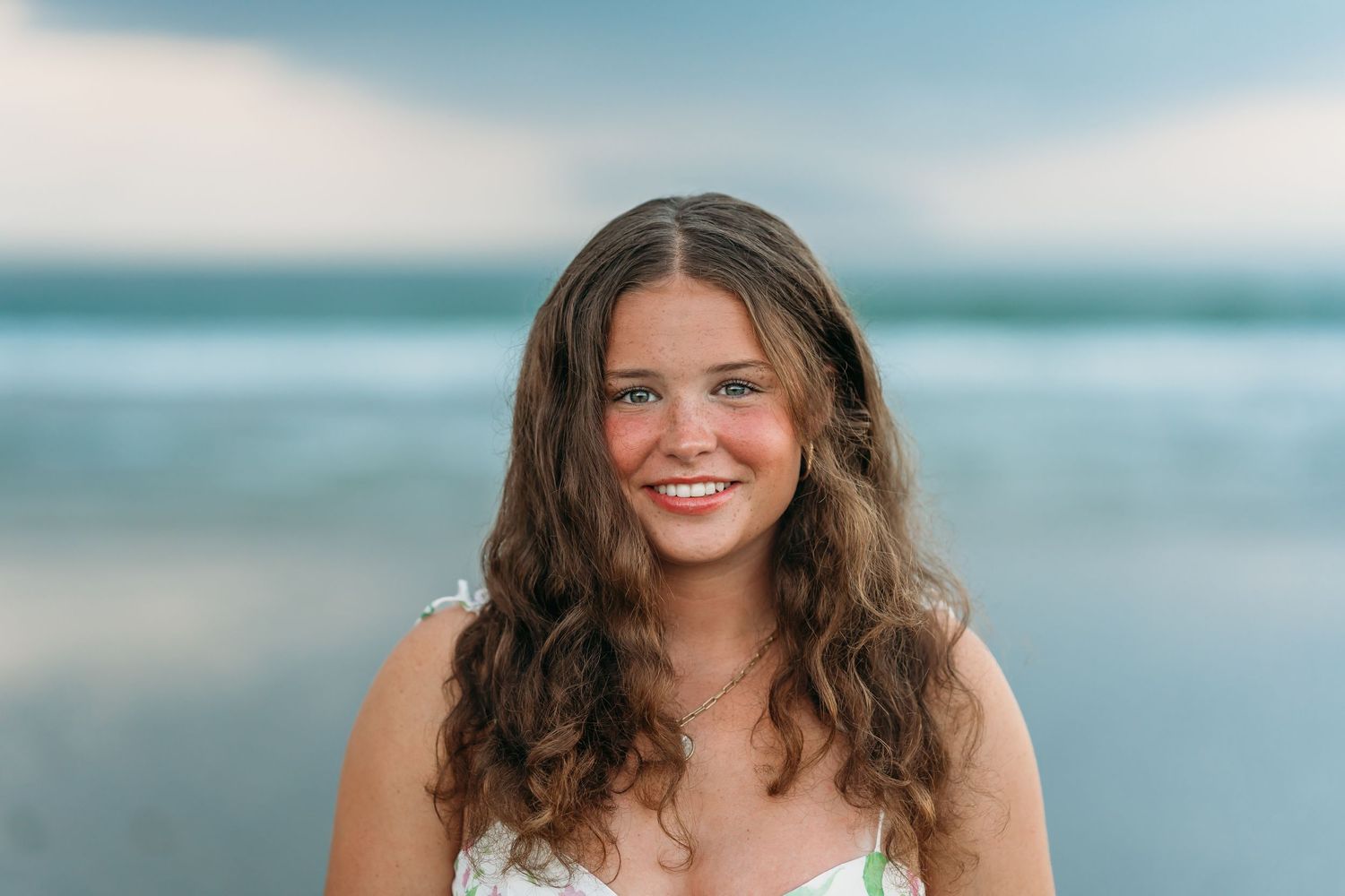 Smiling young woman with long wavy brown hair posing at the beach with ocean in the background.