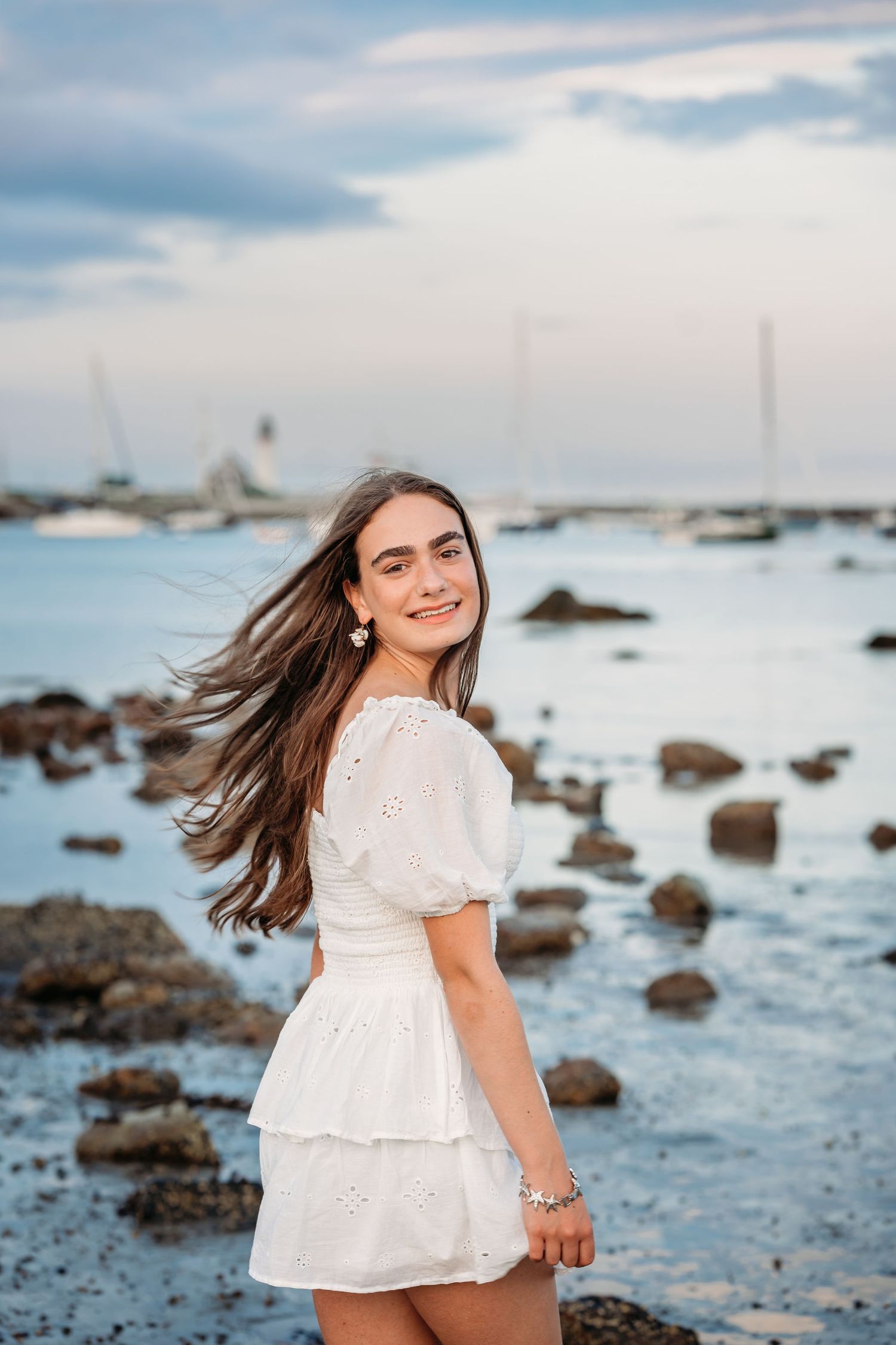 A white dress flows in the breeze at the rocky beach with sailboats and calm waters in the background.