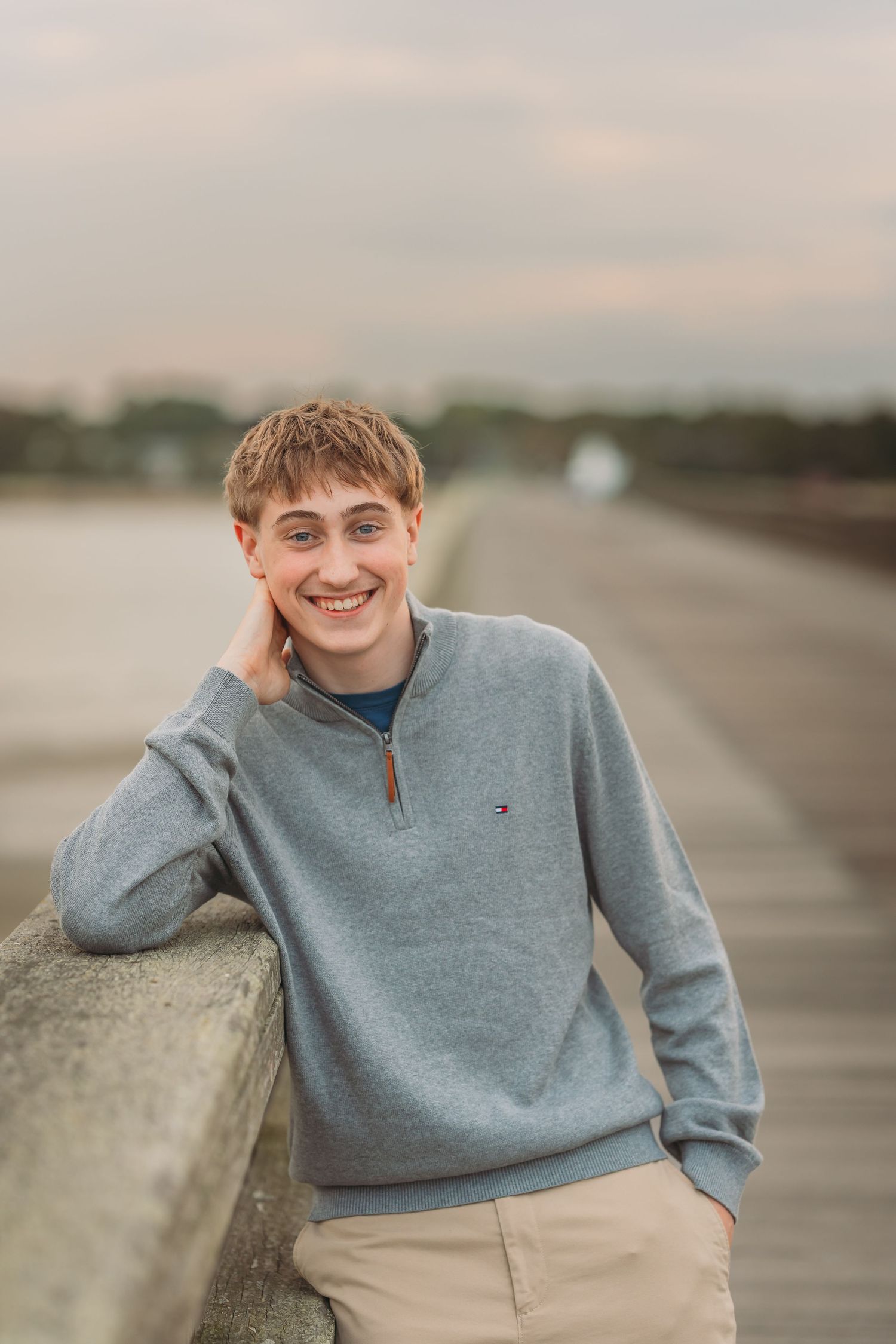 Smiling young man in gray Tommy Hilfiger quarter-zip sweater leaning on a railing outdoors.
