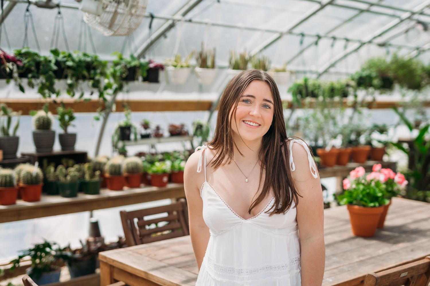 Smiling young woman in white dress standing in a lush greenhouse surrounded by potted plants and cacti.