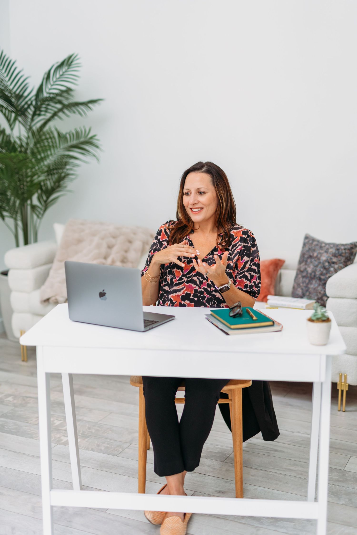 Woman smiling at desk with MacBook laptop, books, and plant in bright home office workspace.
