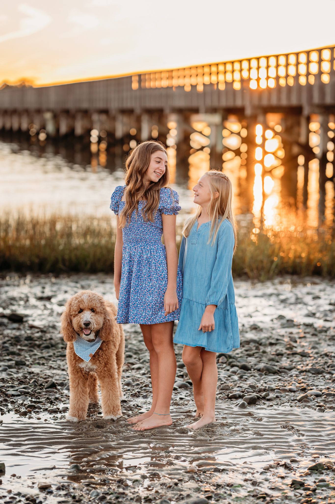 Two girls in summer dresses stand with their goldendoodle dog by a pier during golden hour sunset.