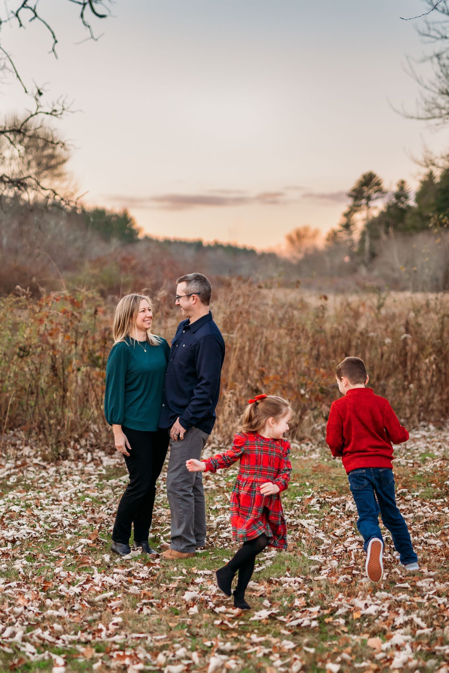 A family in matching winter outfits walks through a snowy field at sunset with tall grass and trees in the background.
