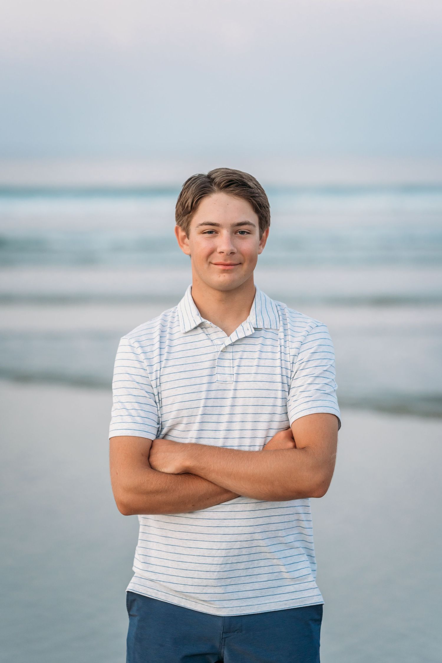 Teen boy with crossed arms smiling on beach, wearing striped polo shirt with ocean waves in background.