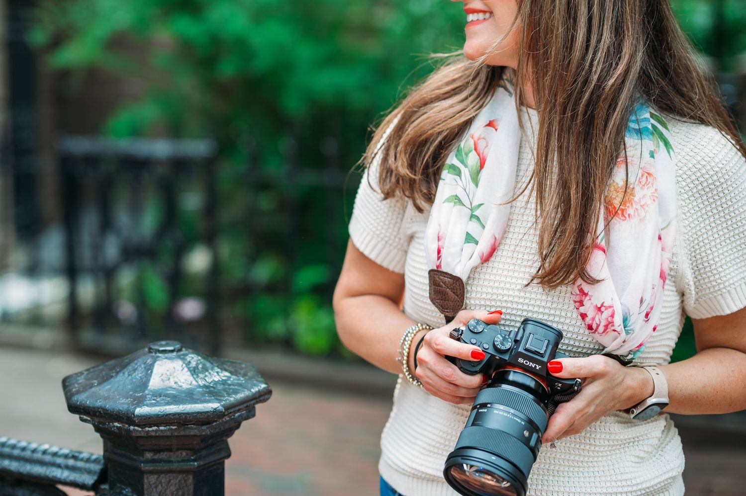 Photographer wearing floral scarf and white top holds professional camera while smiling outdoors.