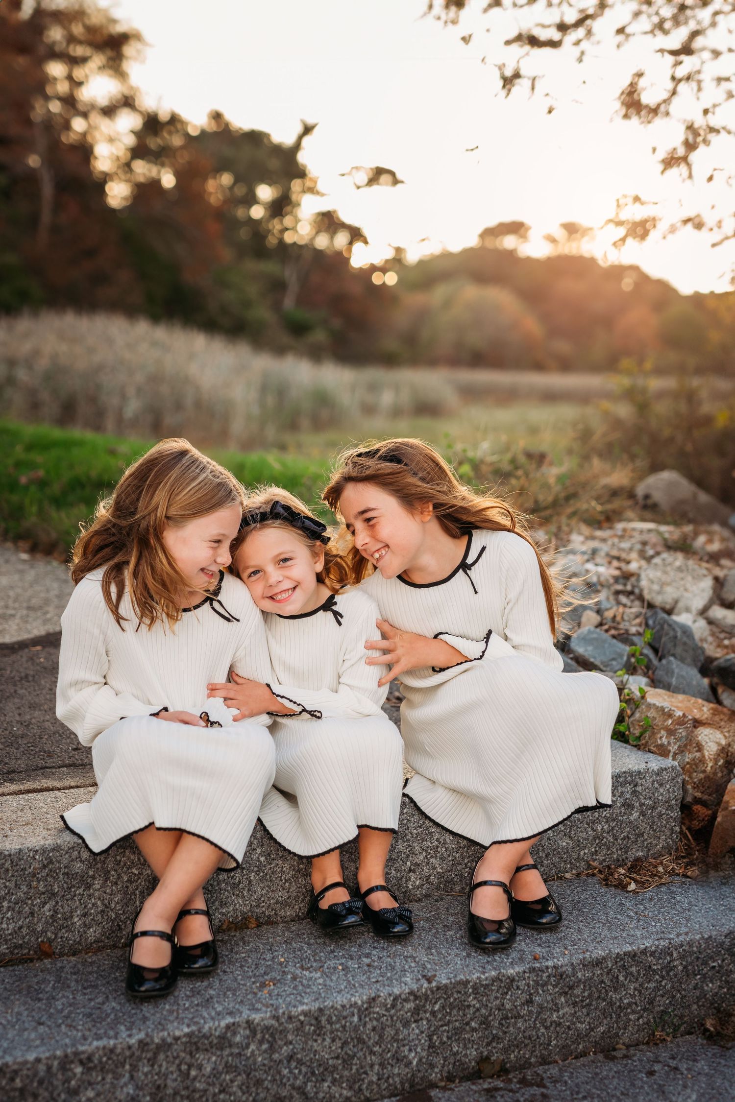 Three people in white dresses sit together on a stone wall at sunset, sharing joyful moments and laughter.