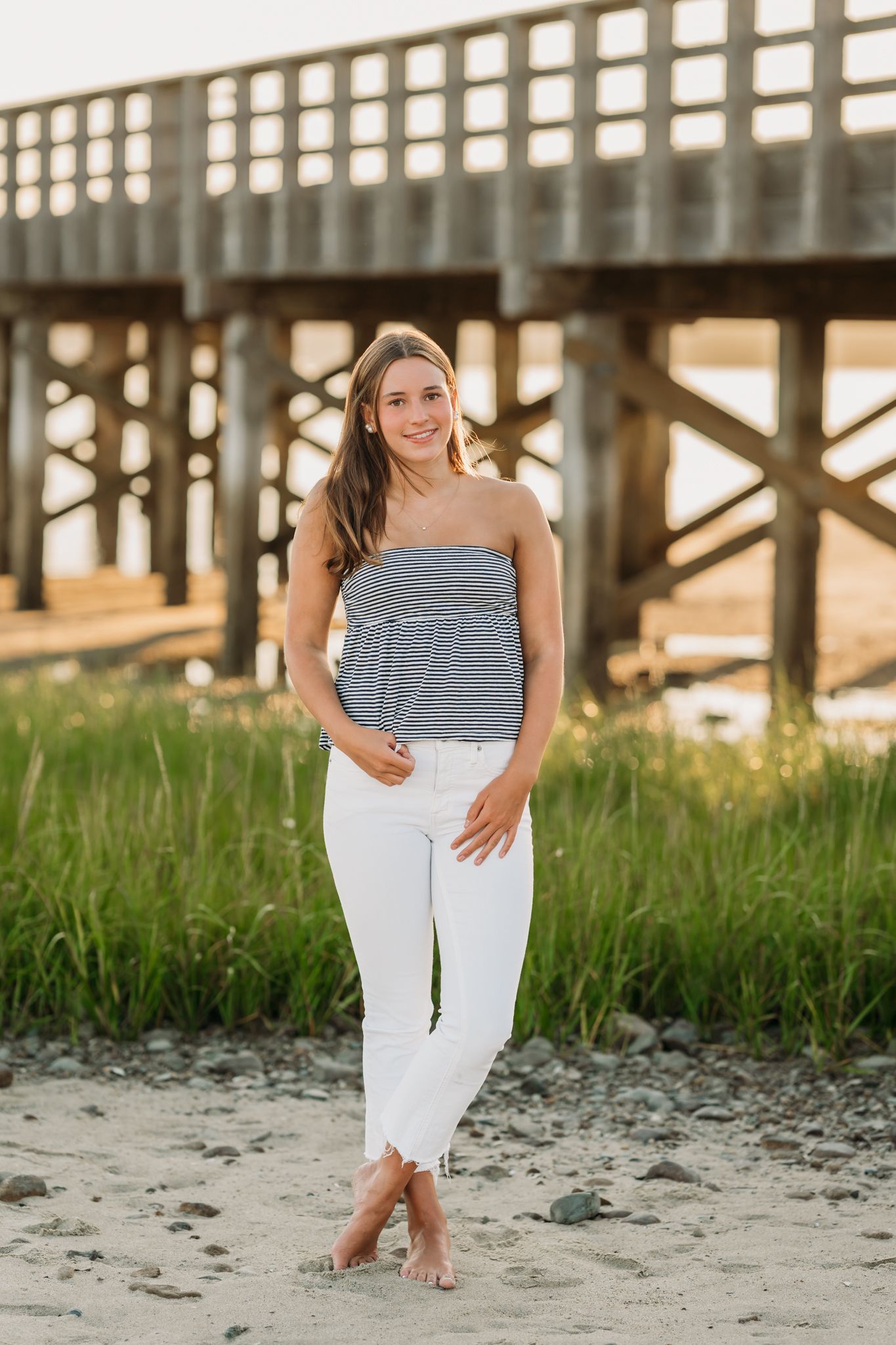 Young woman in white jeans and striped tube top poses barefoot on sandy beach near wooden pier.