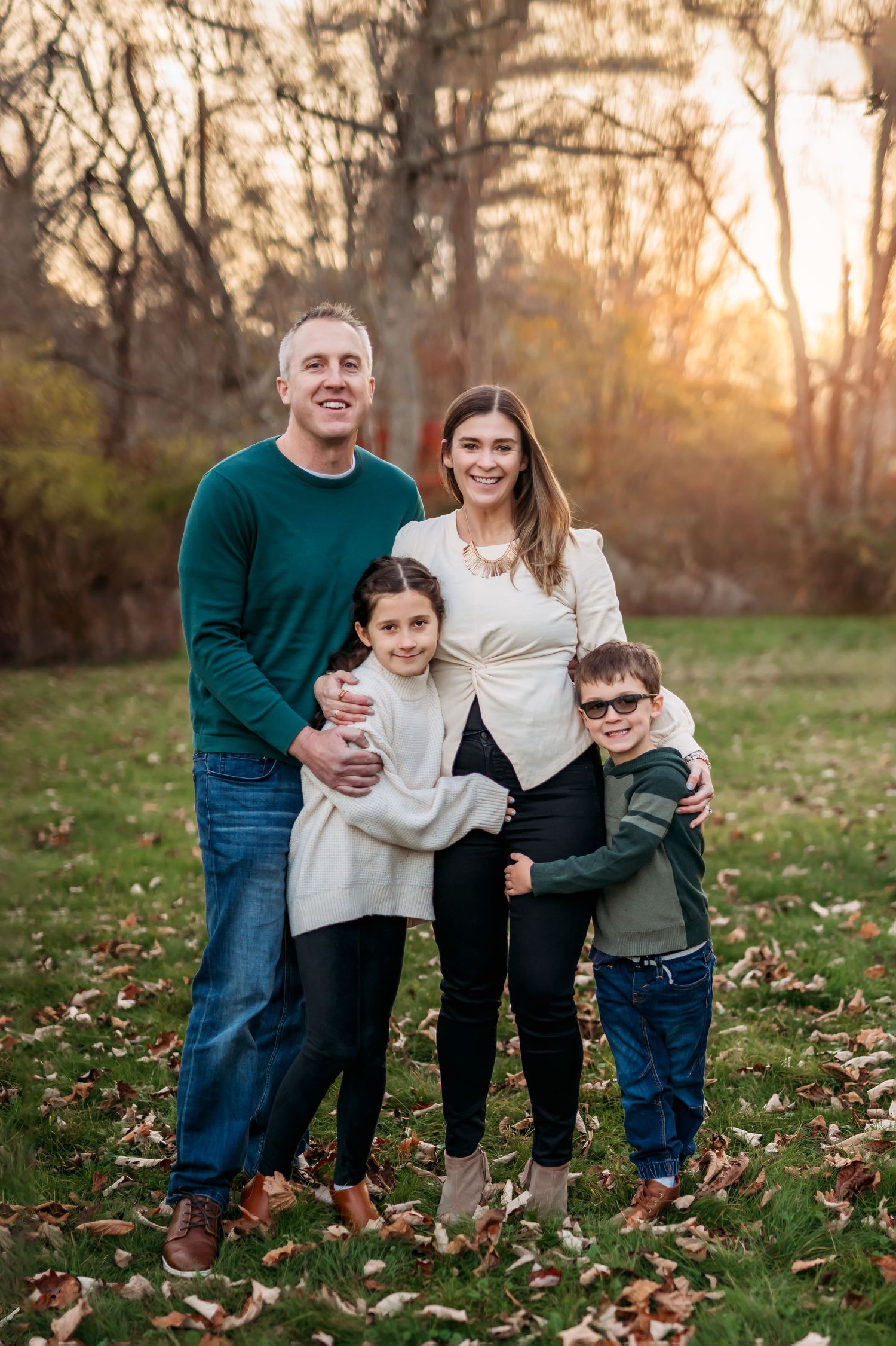 Family wearing coordinated white and green outfits poses together on autumn grass covered with fallen leaves at sunset.